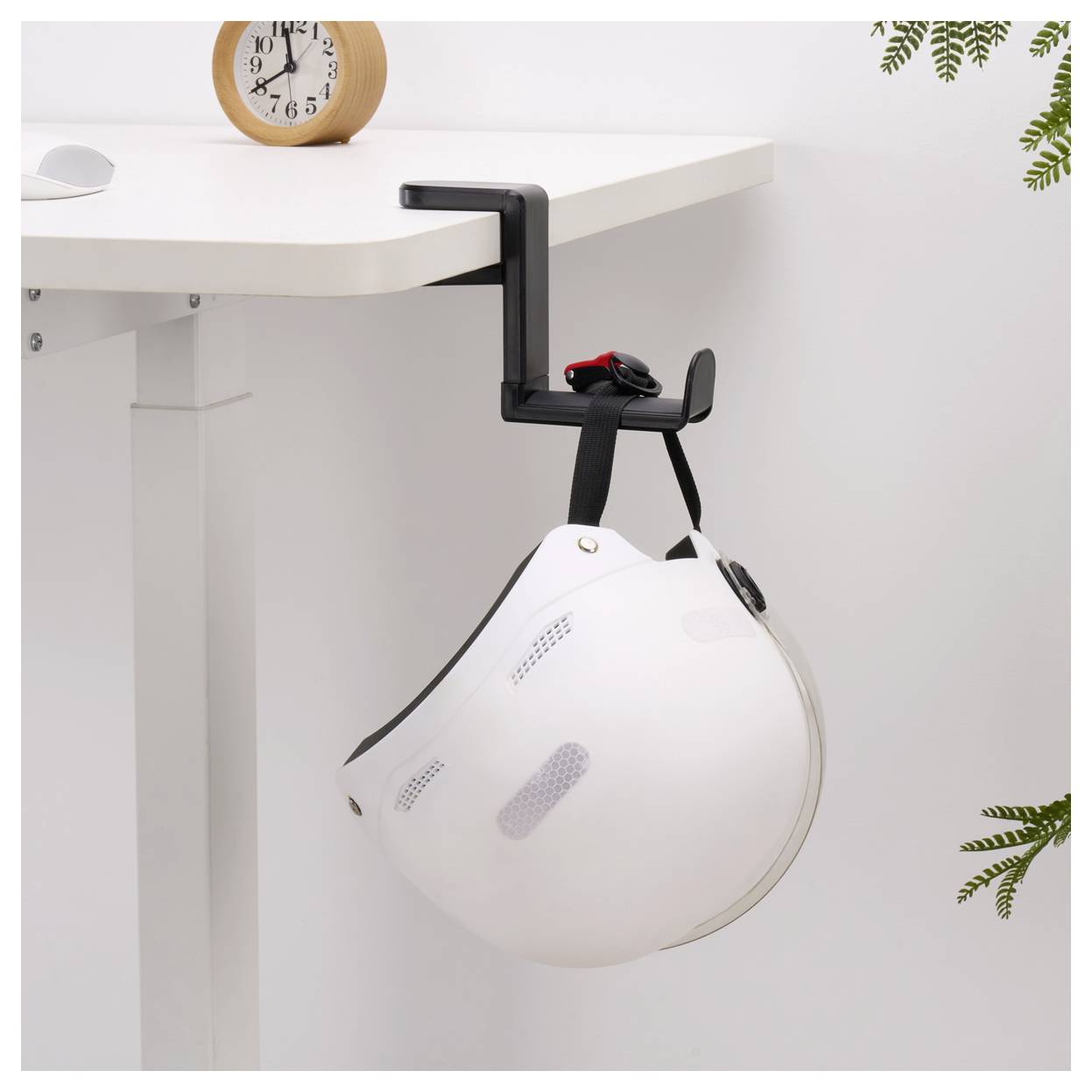 'A white protective helmet hanging on a table by a black clamp hook, with a small wooden clock on the table and plant leaves nearby.'