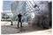 A worker uses a high-pressure power washer on a tall modern glass and stone building, with water and cleaning hose visible.