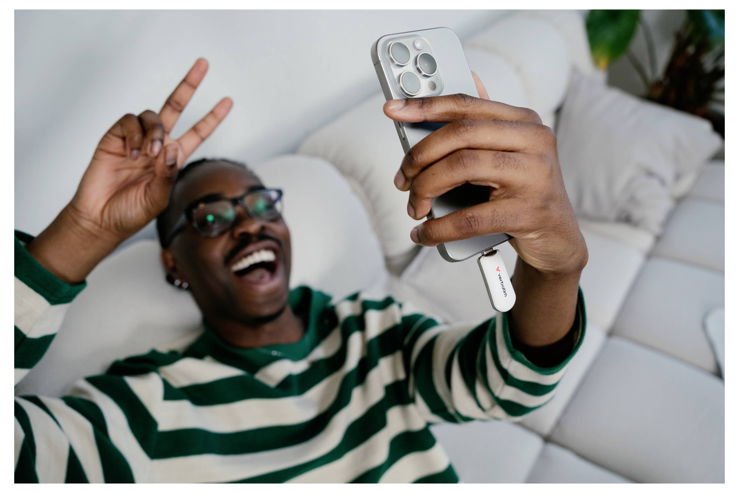 Smiling man in a striped sweater takes a selfie on a couch, flashing a peace sign and holding a phone with a white accessory attached.