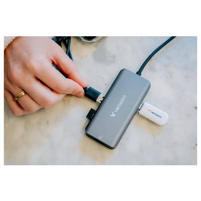 Hand plugs a USB cable into a gray Verbatim hub with a white USB drive attached on a marble surface.