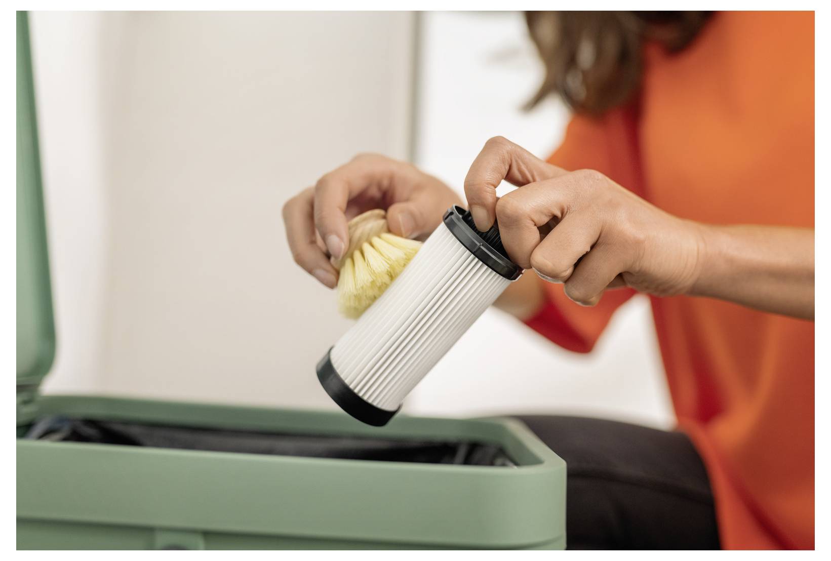 A person in an orange shirt holds a cylindrical air filter and a crumpled cloth, demonstrating how to change the filter in a green device.