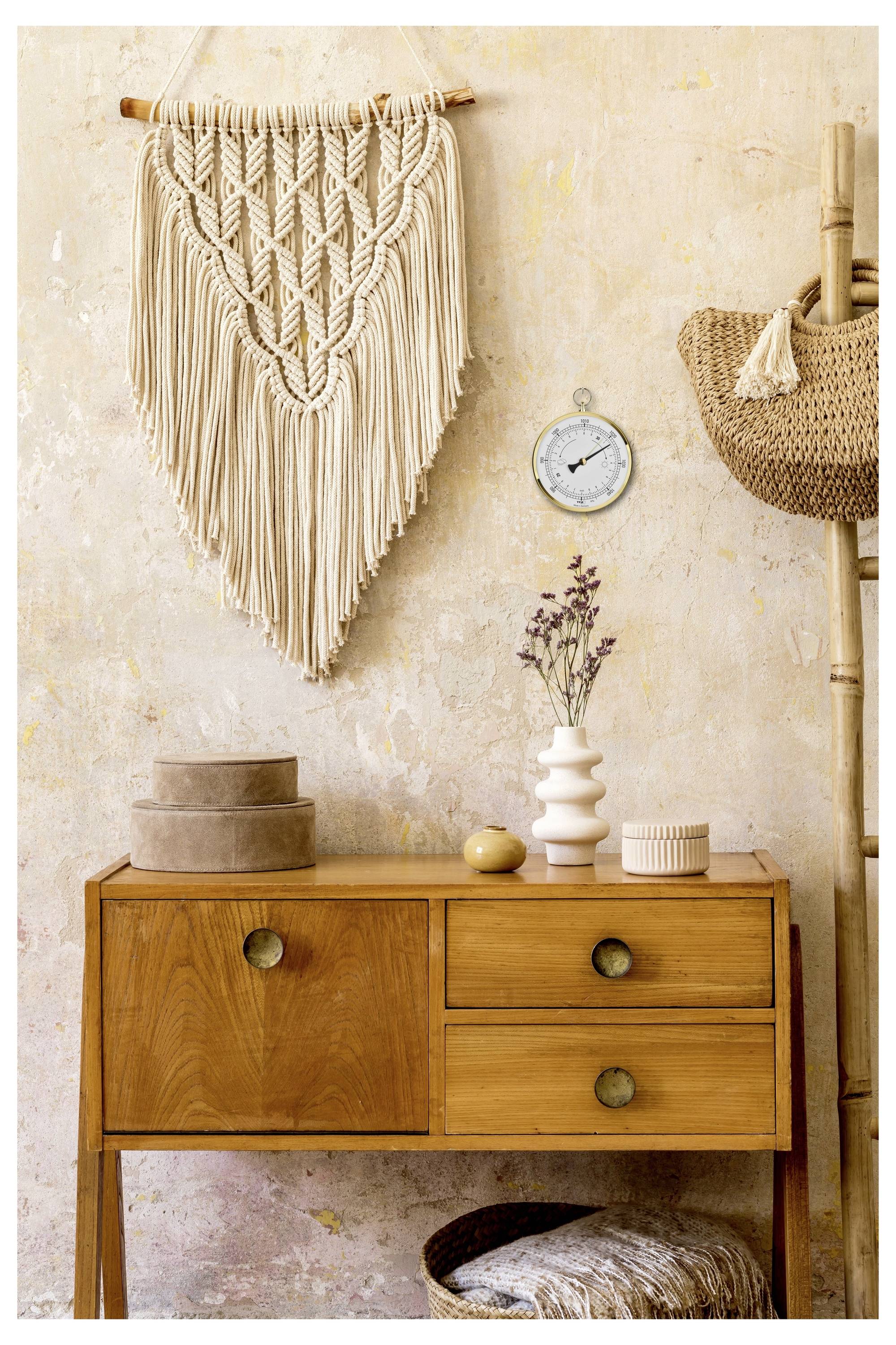 A wooden table with drawers holds decorative objects: a vase with dried flowers, a stack of round boxes, and a wicker basket. Above, a fringed macramé wall hanging and a small round clock.