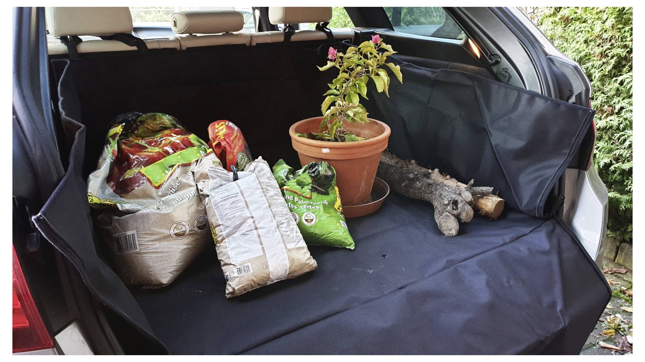 'Several bags of soil, a potted plant with pink flowers, and tree branches are arranged in the open trunk of a car.'