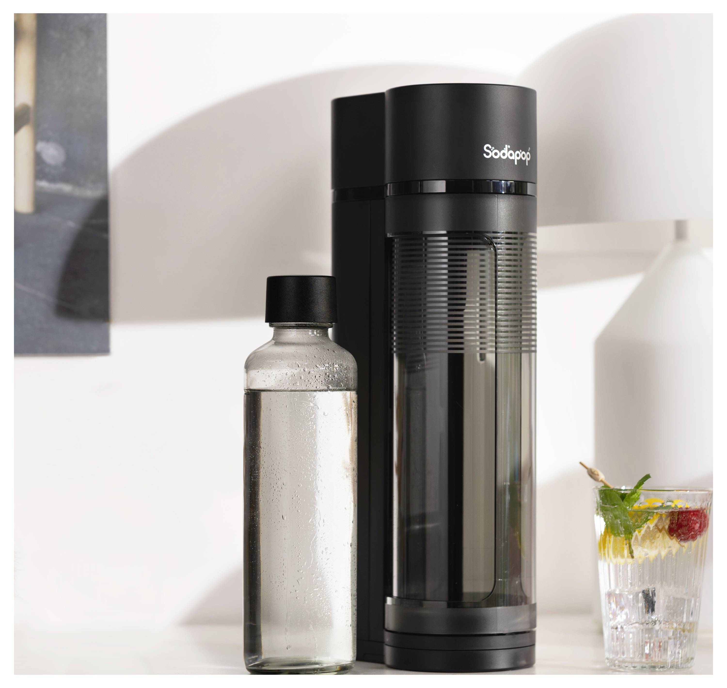 A black soda maker stands on a counter beside a clear bottle filled with water and a glass of sparkling drink with mint and fruit garnish.