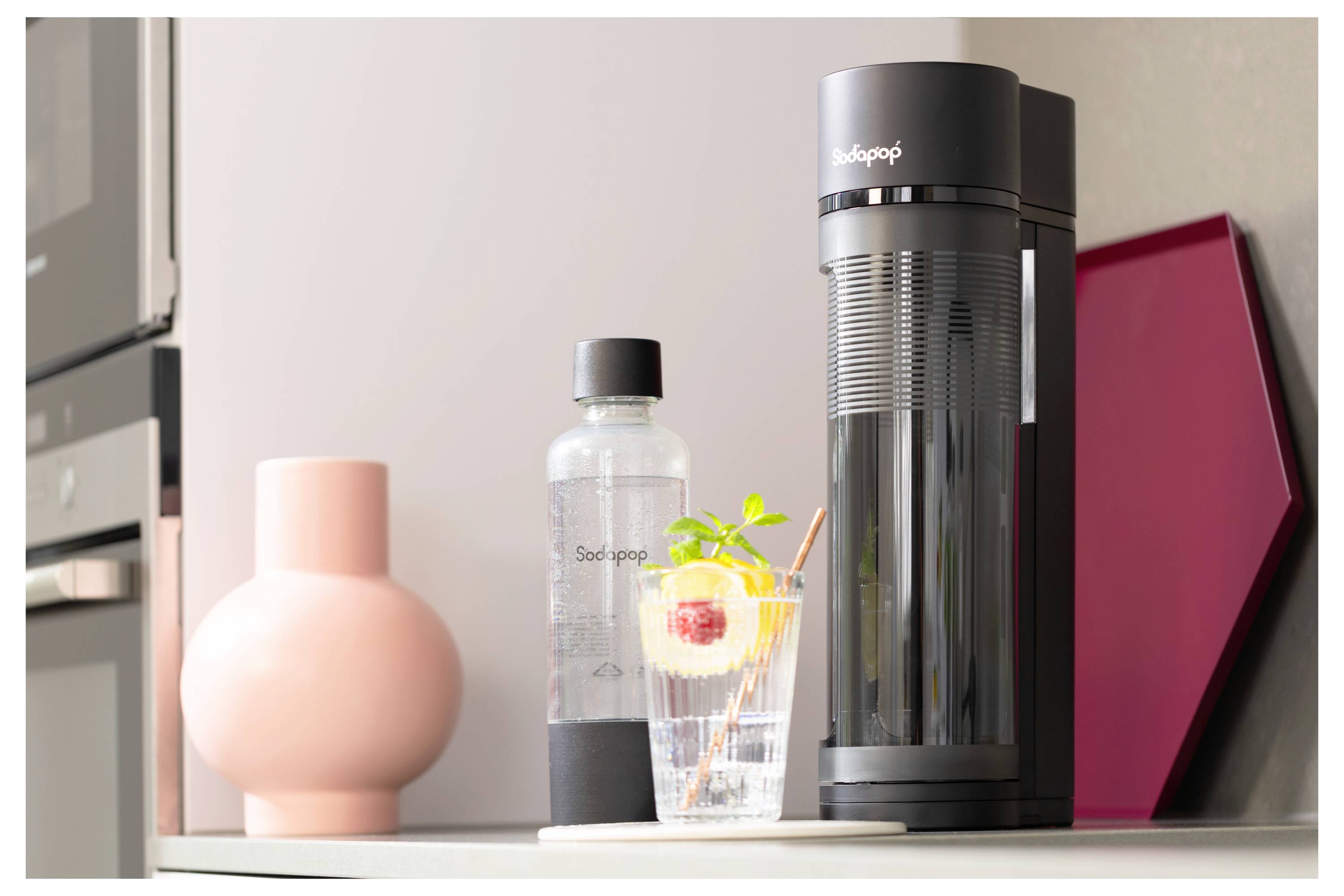 A black soda maker on a kitchen counter next to a bottle and a glass with iced fruit water.