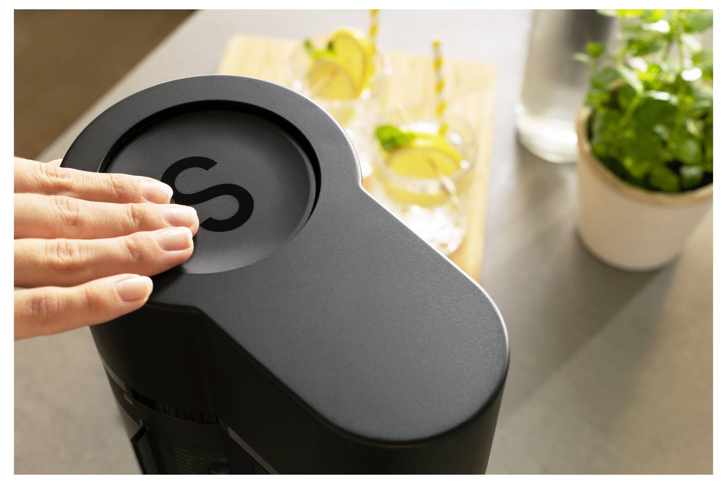A hand presses the top button of a modern kitchen appliance with two drinks and a plant in the background, suggesting beverage preparation.
