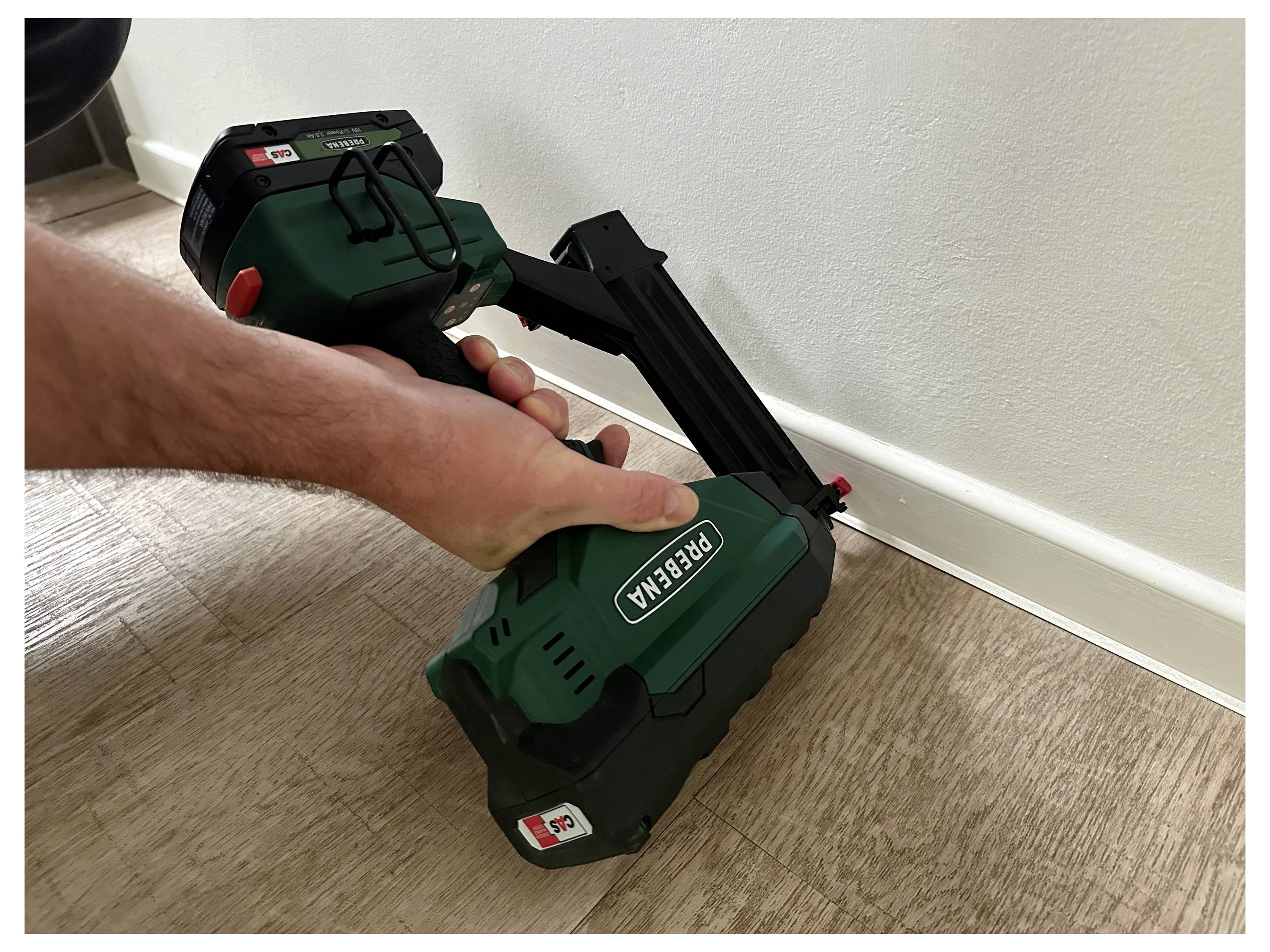A person uses a green nail gun labeled 'PRESA' to install white baseboards onto a wood floor.