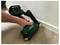 A person uses a green nail gun labeled 'PRESA' to install white baseboards onto a wood floor.