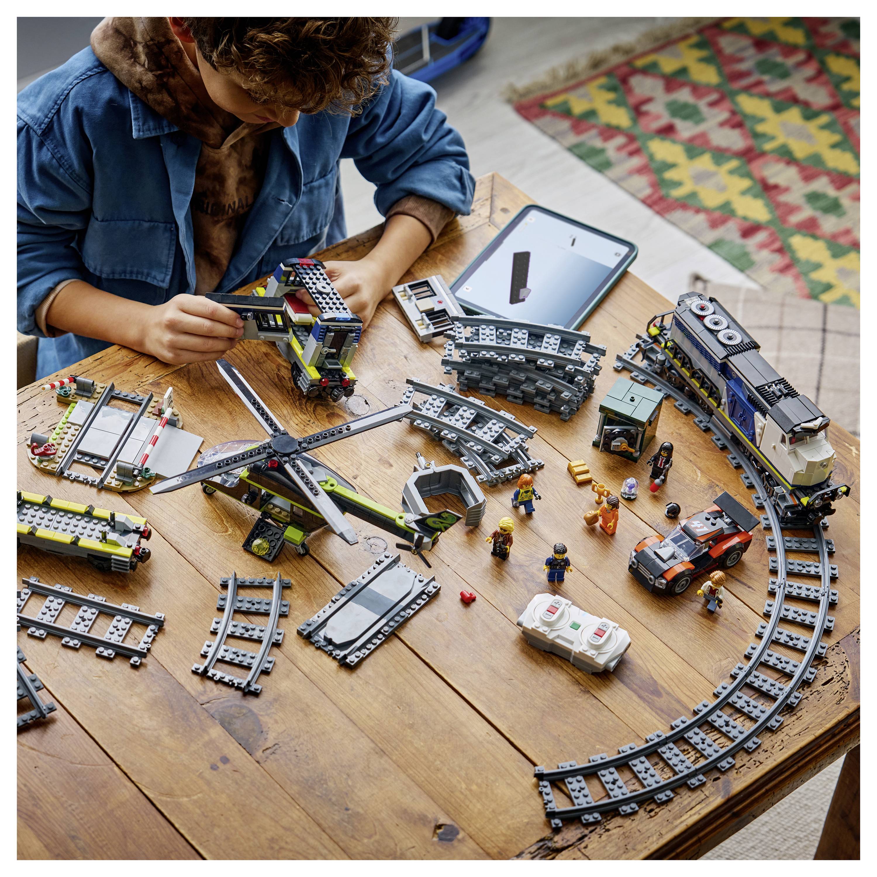 A child plays with a train set and various toy vehicles on a wooden table, surrounded by curved and straight train tracks and a tablet.