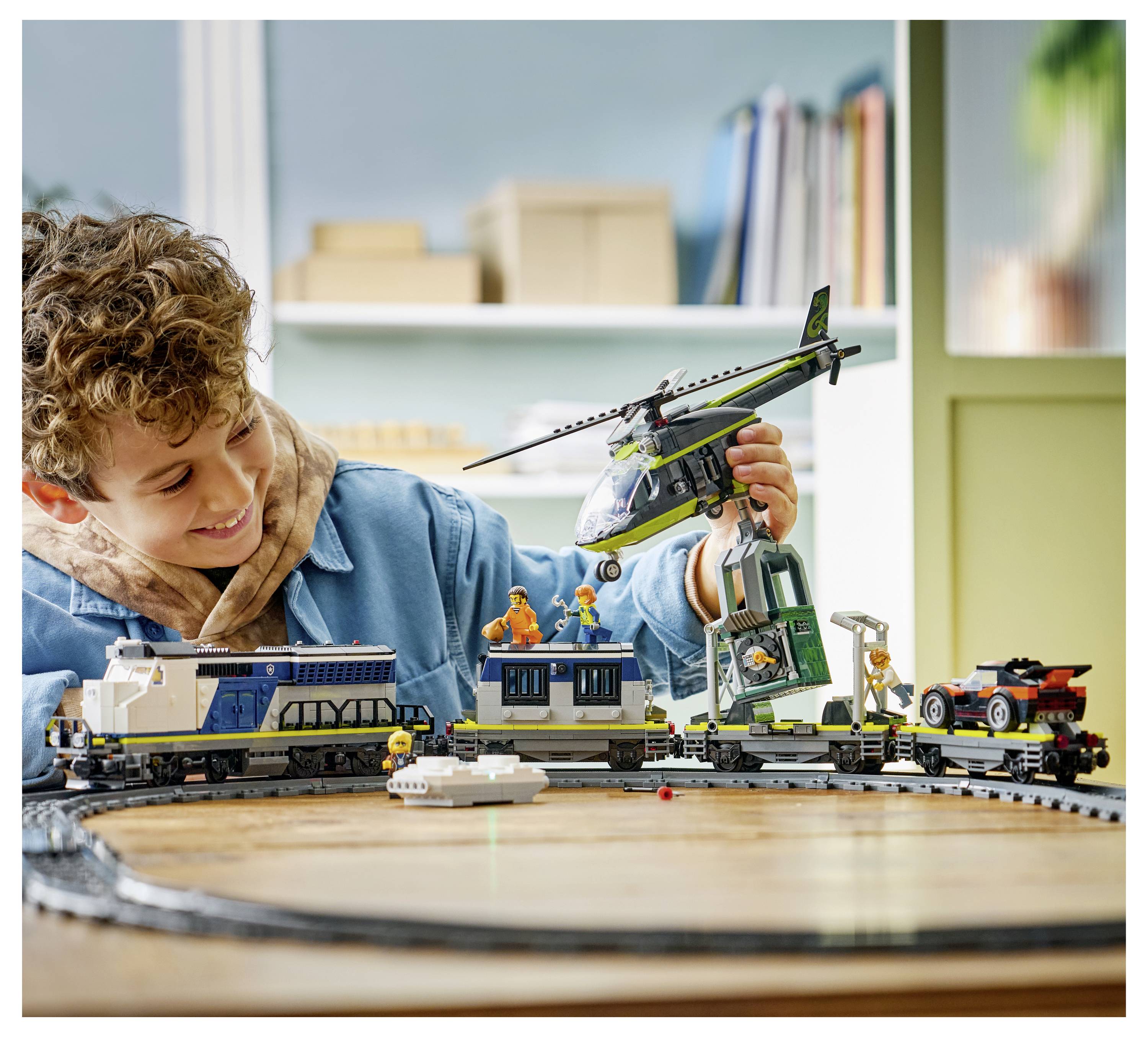 A child plays with a train and helicopter toy set on a circular track. The backdrop includes shelves with books and boxes.