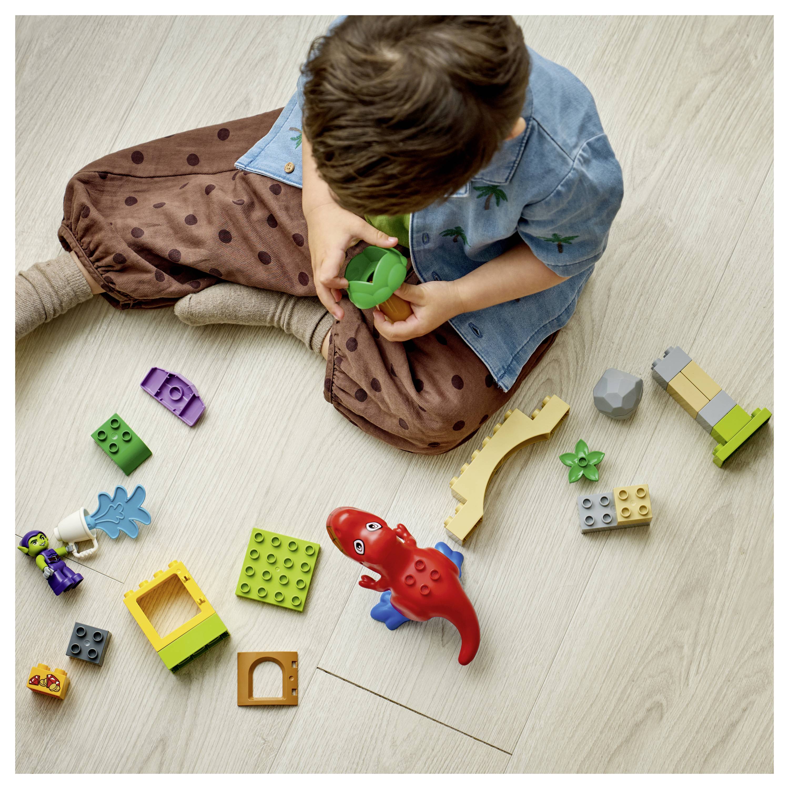 A child is sitting on a wooden floor, playing with colorful building blocks and a red dinosaur toy.