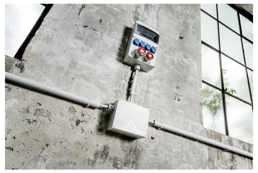 Industrial electrical boxes and conduit mounted on a concrete wall beside tall window panes.