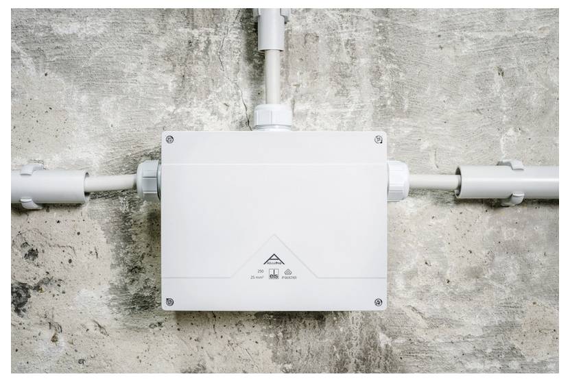 White electrical junction box mounted on a concrete wall with pipes entering from the top and both sides.