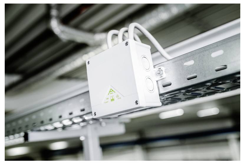 White electrical control box mounted on a metal cable tray, with conduit wires attached in an industrial ceiling area.
