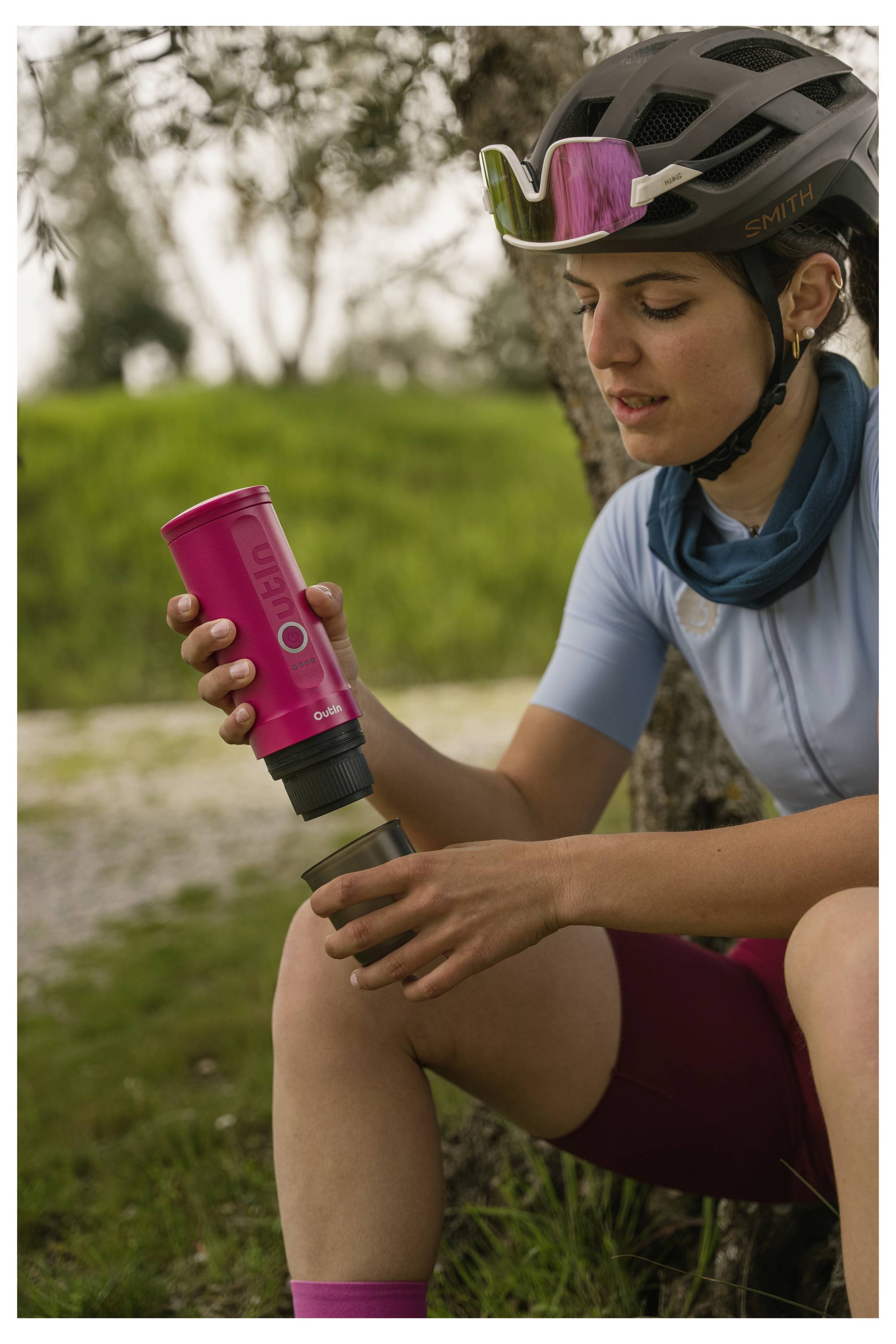A person wearing cycling gear, including a helmet and sunglasses, sits outdoors, pouring a drink from a pink thermos into a cup on their knee.