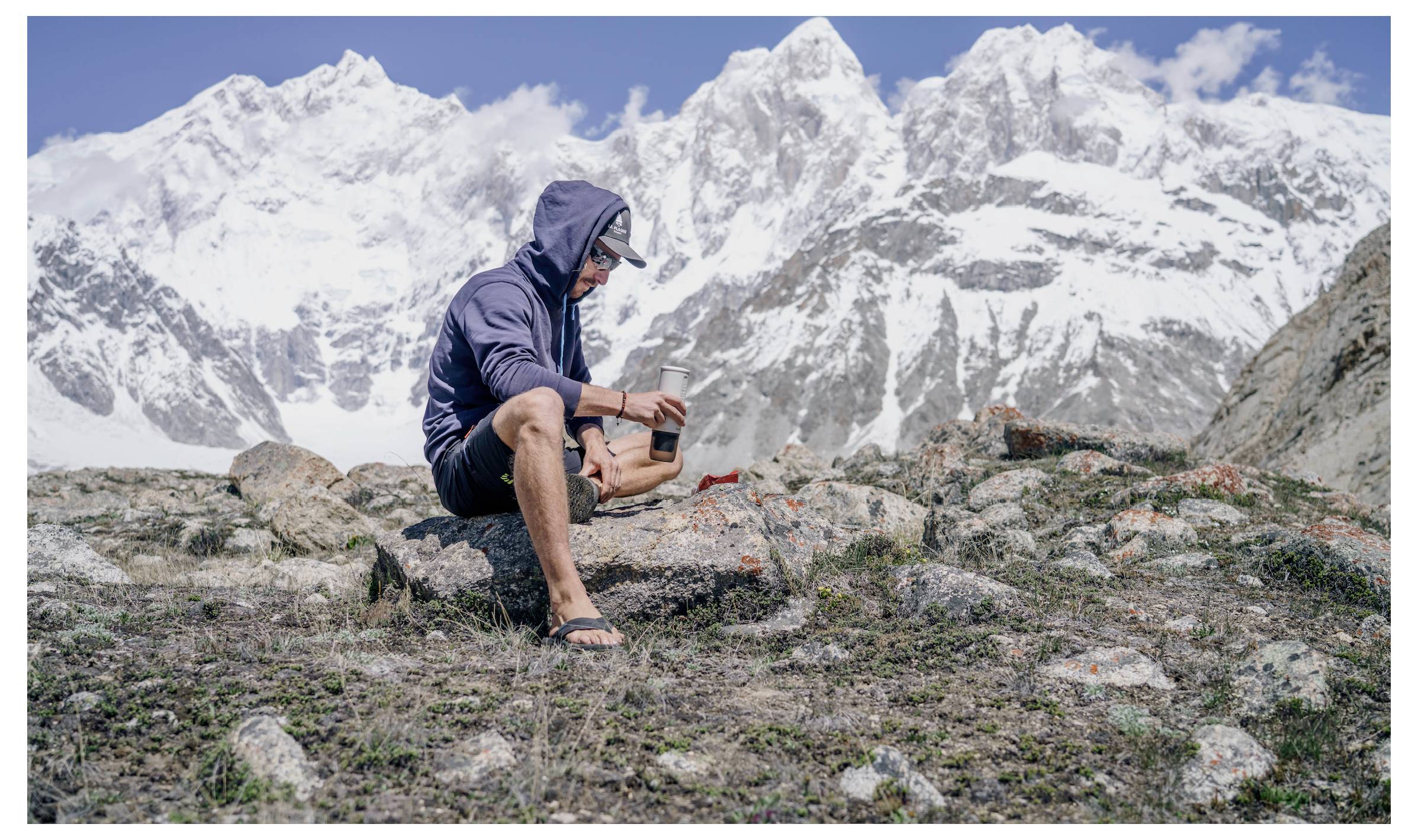 Person in casual outdoor attire sits on rocky ground using smartphone, with snow-capped mountains visible in the background.