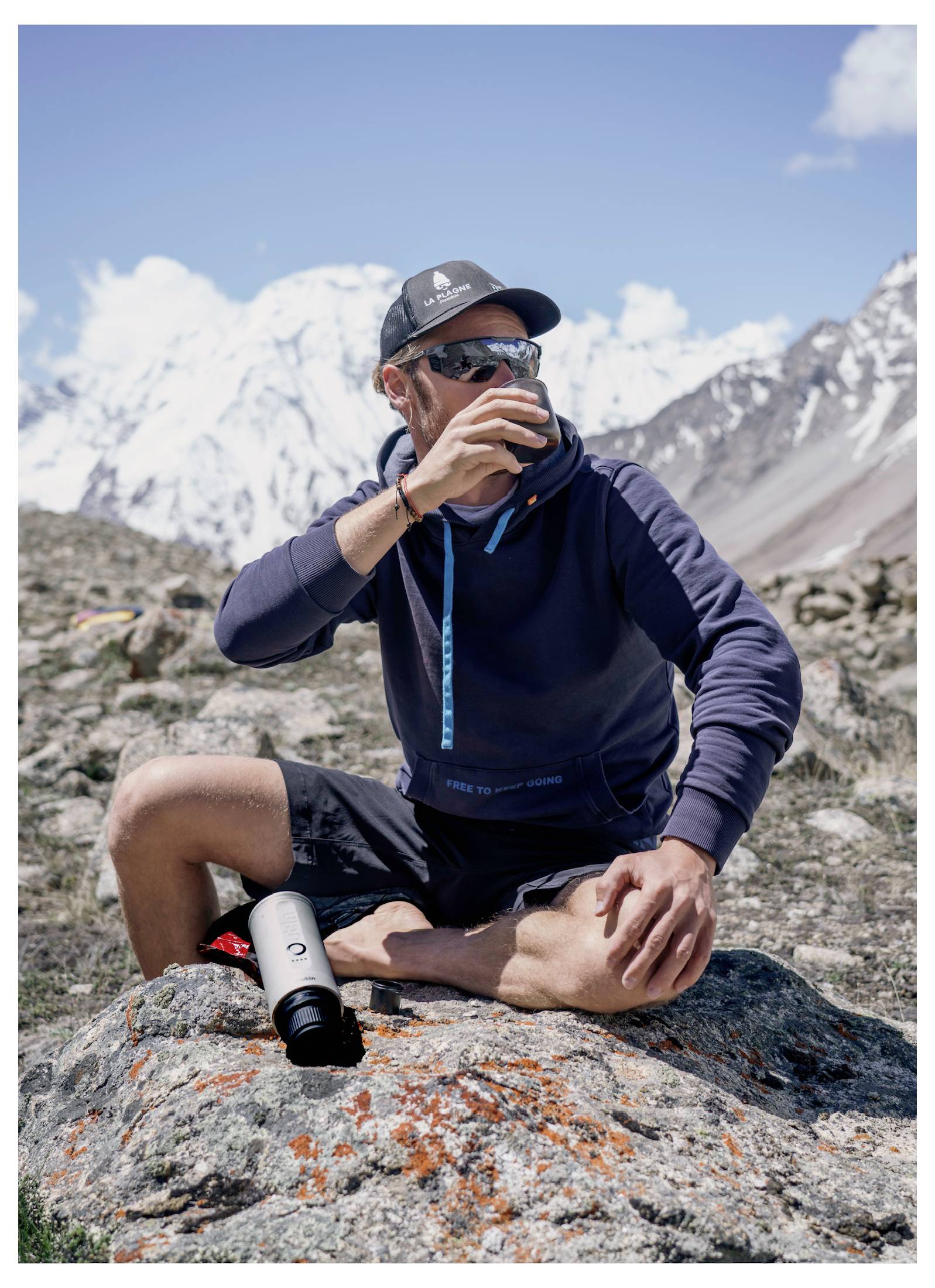A person in outdoor attire sits on a rock in a mountainous area, drinking from a bottle. Snow-capped peaks are visible in the background.