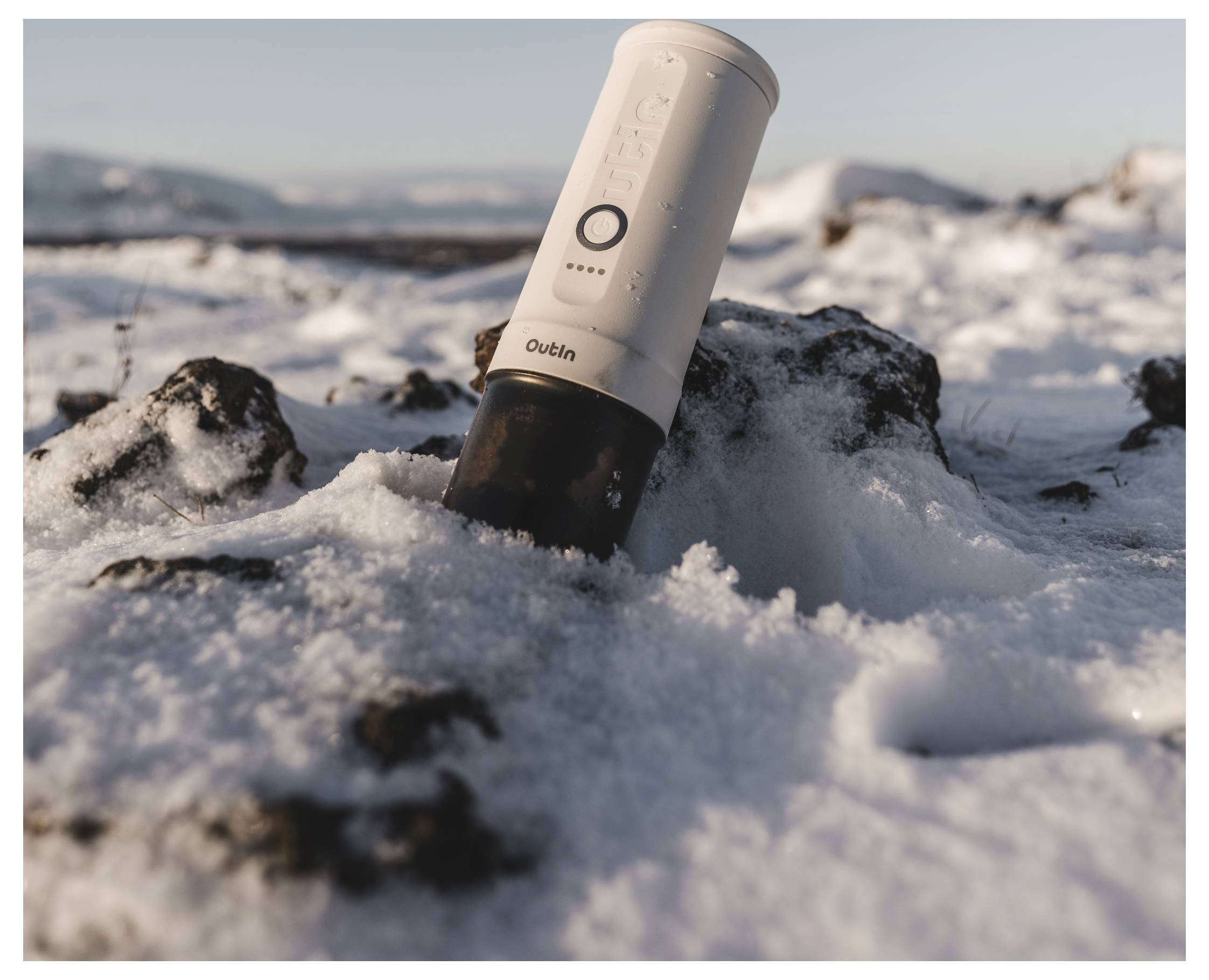 'A portable coffee grinder partially buried in snow on a rocky landscape, with distant mountains in the background under a clear sky.'