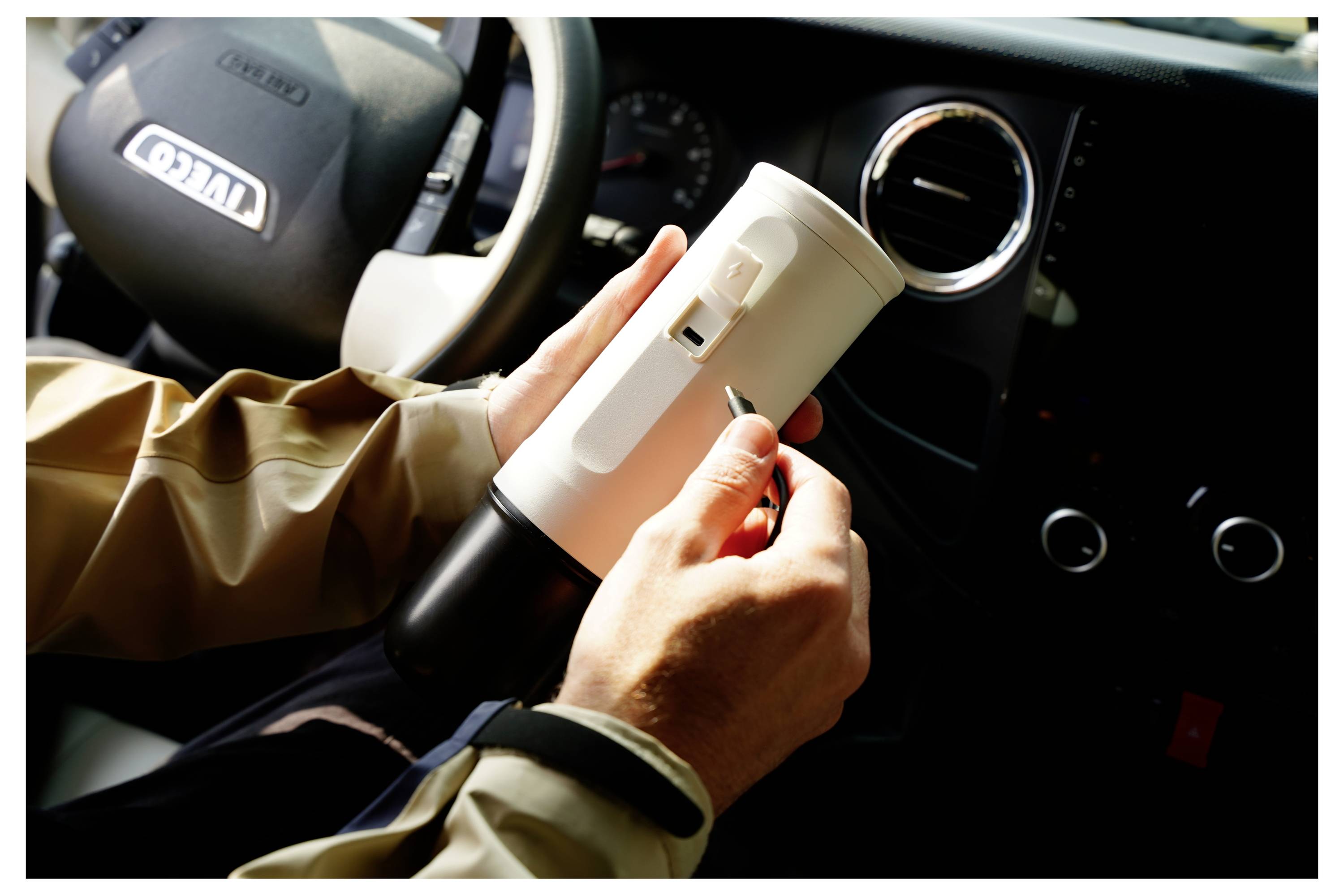 A person in a vehicle holding a white cylindrical object, resembling a small appliance, adjusting a switch on its side. Dashboard visible.