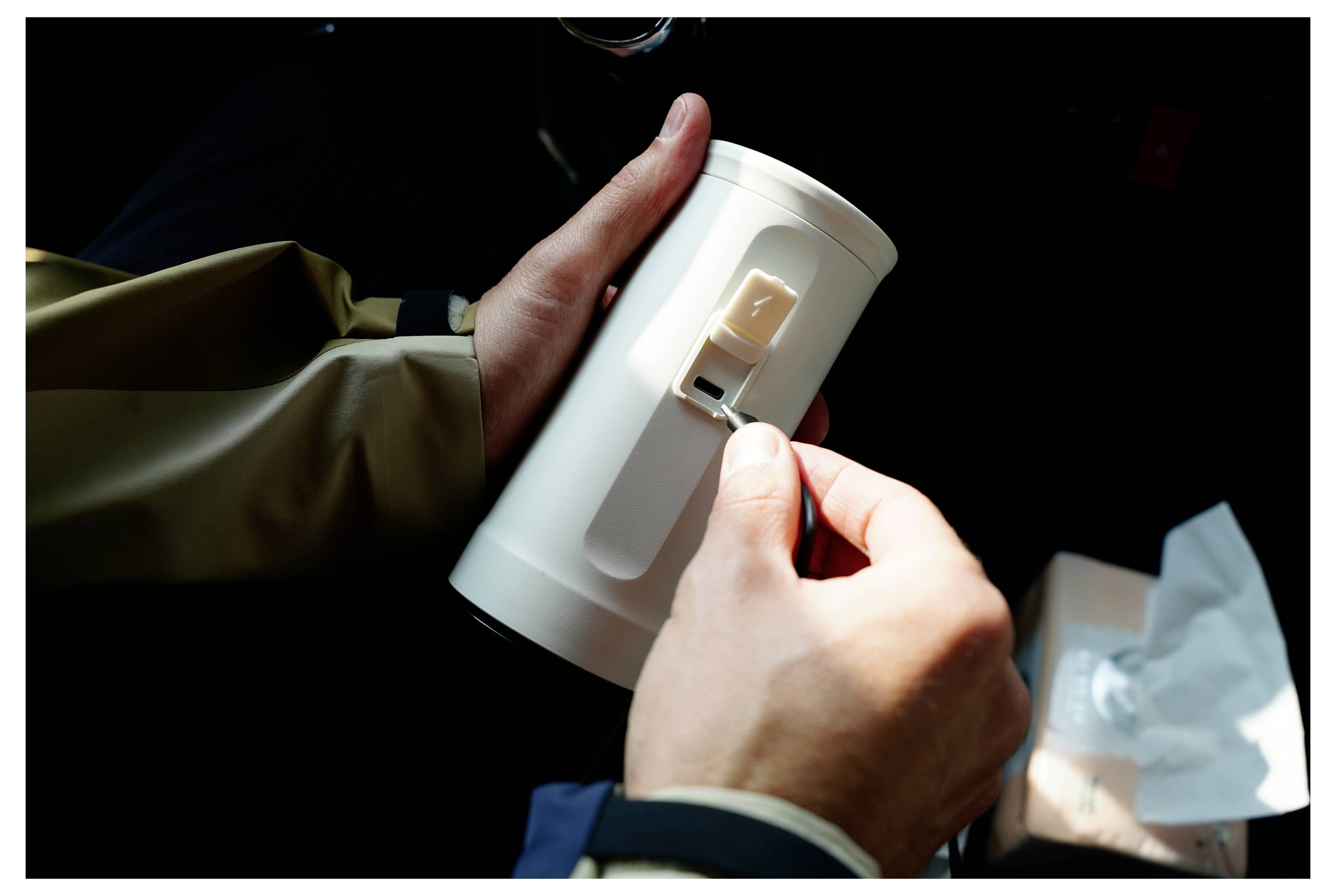 A person adjusts the lid on a cylindrical container with a clip, seated inside a vehicle. Light shines on the hands and container.