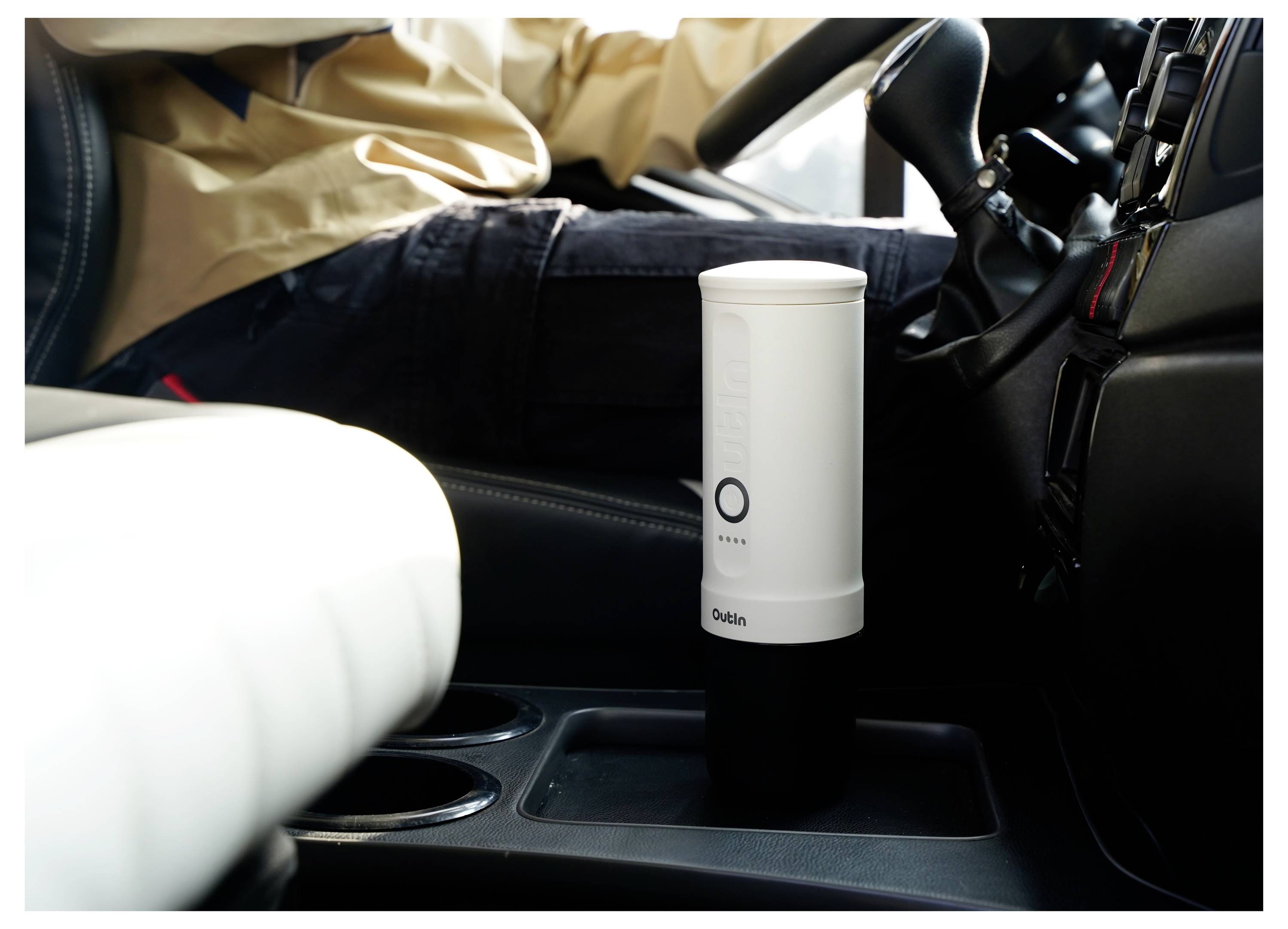 A person driving a car while a white travel mug sits in the cup holder beside the driver's seat.