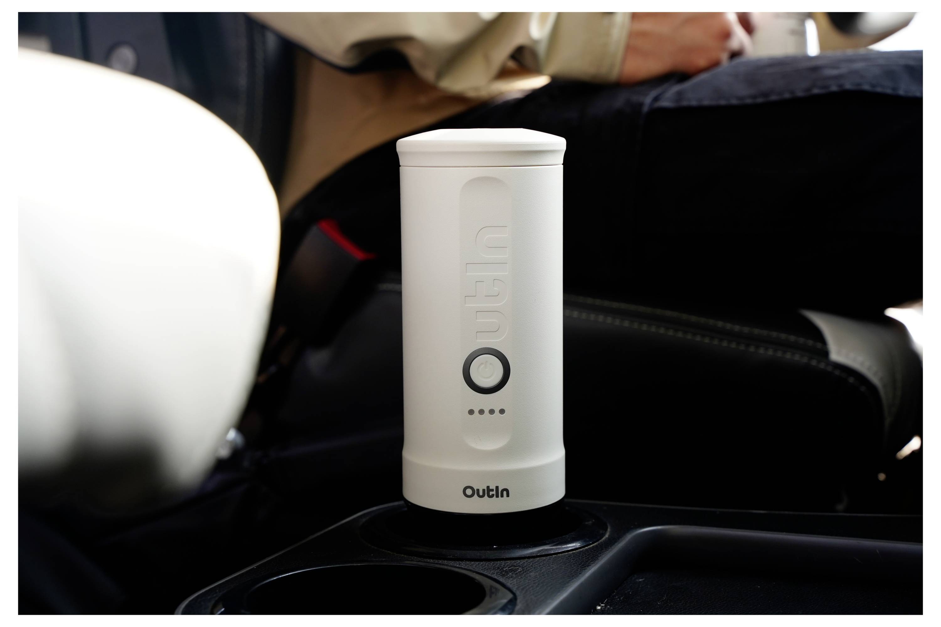 A portable white car kettle labeled 'Outin' is placed in a vehicle's cup holder, with a person sitting nearby.