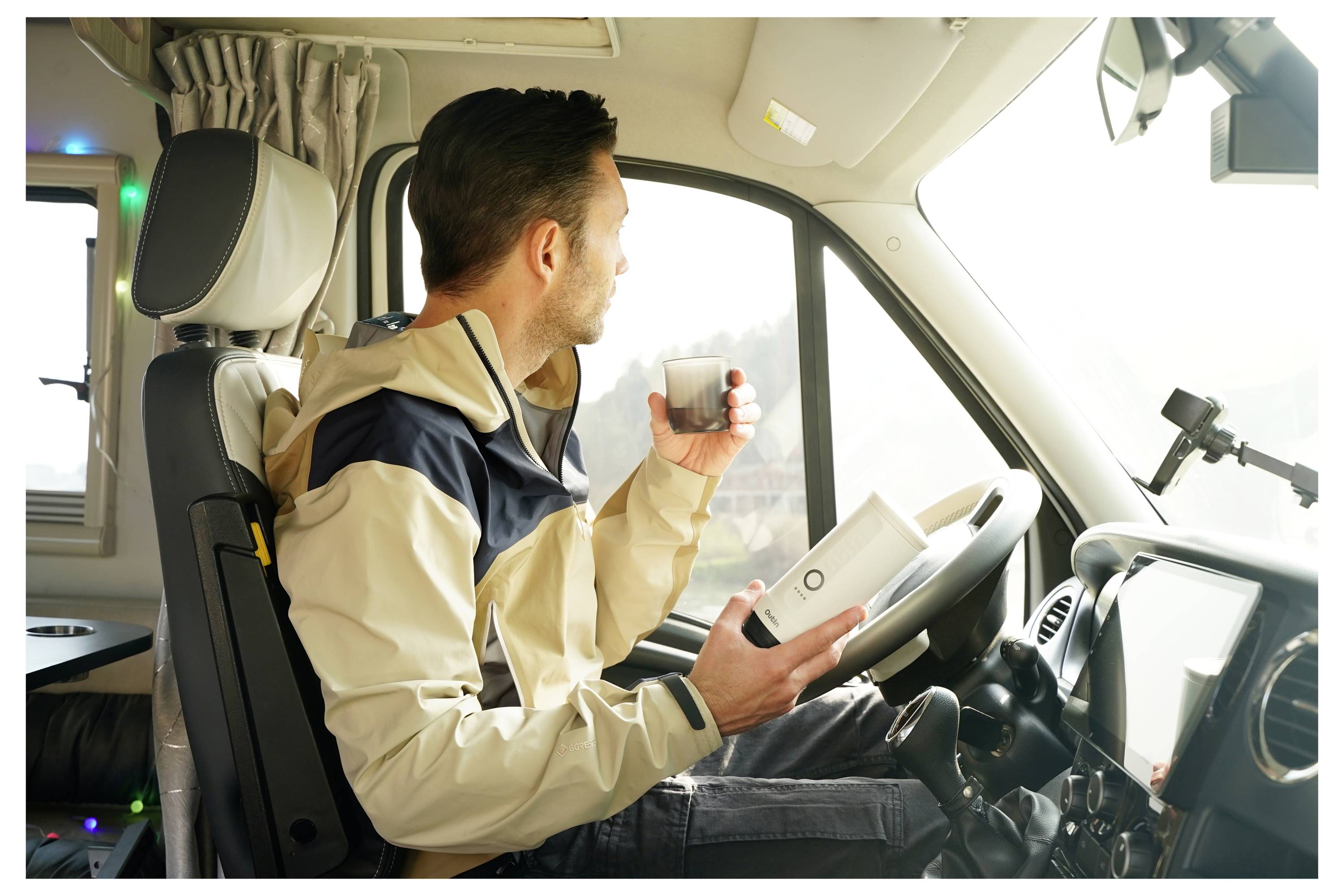 A man in a beige and navy jacket holds a mug and a phone while sitting in the driver's seat of a parked camper van, looking outside.