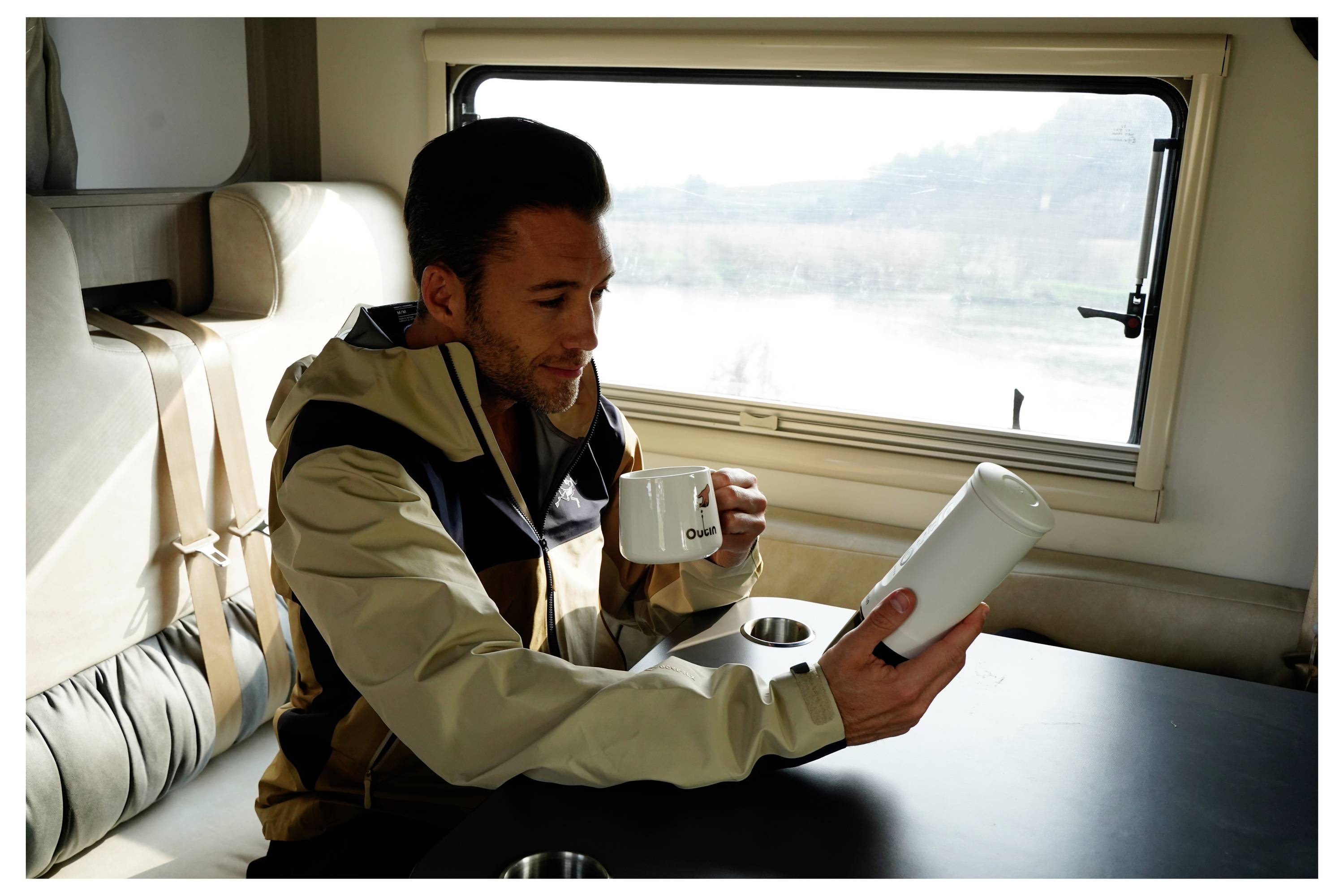 A person in a cozy setting, sitting in a well-lit space while holding a cup and examining a cylindrical object. A large window shows a scenic view.