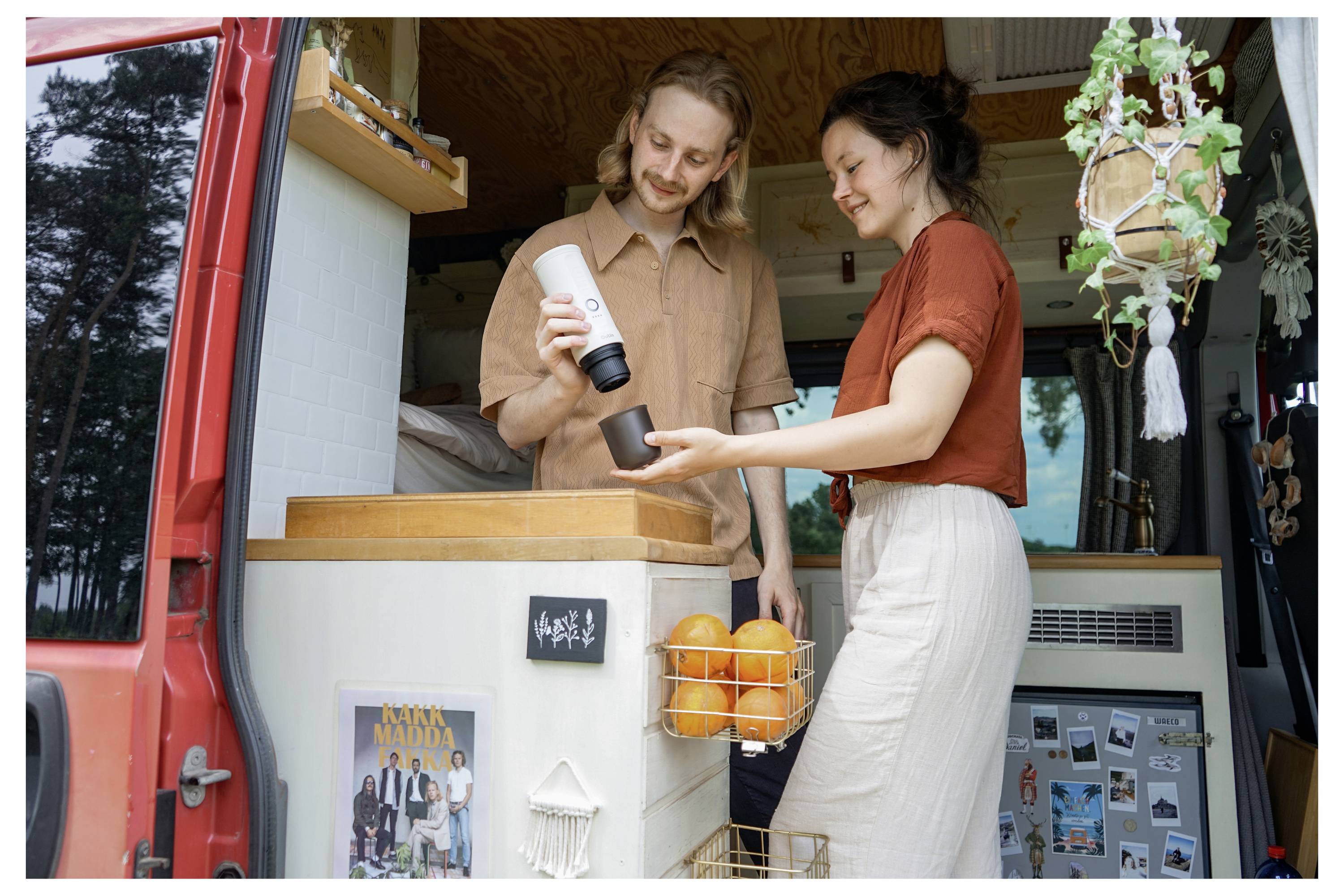 A man and woman prepare a drink in a van converted into a cozy living space, surrounded by plants and personal items, near a forest.