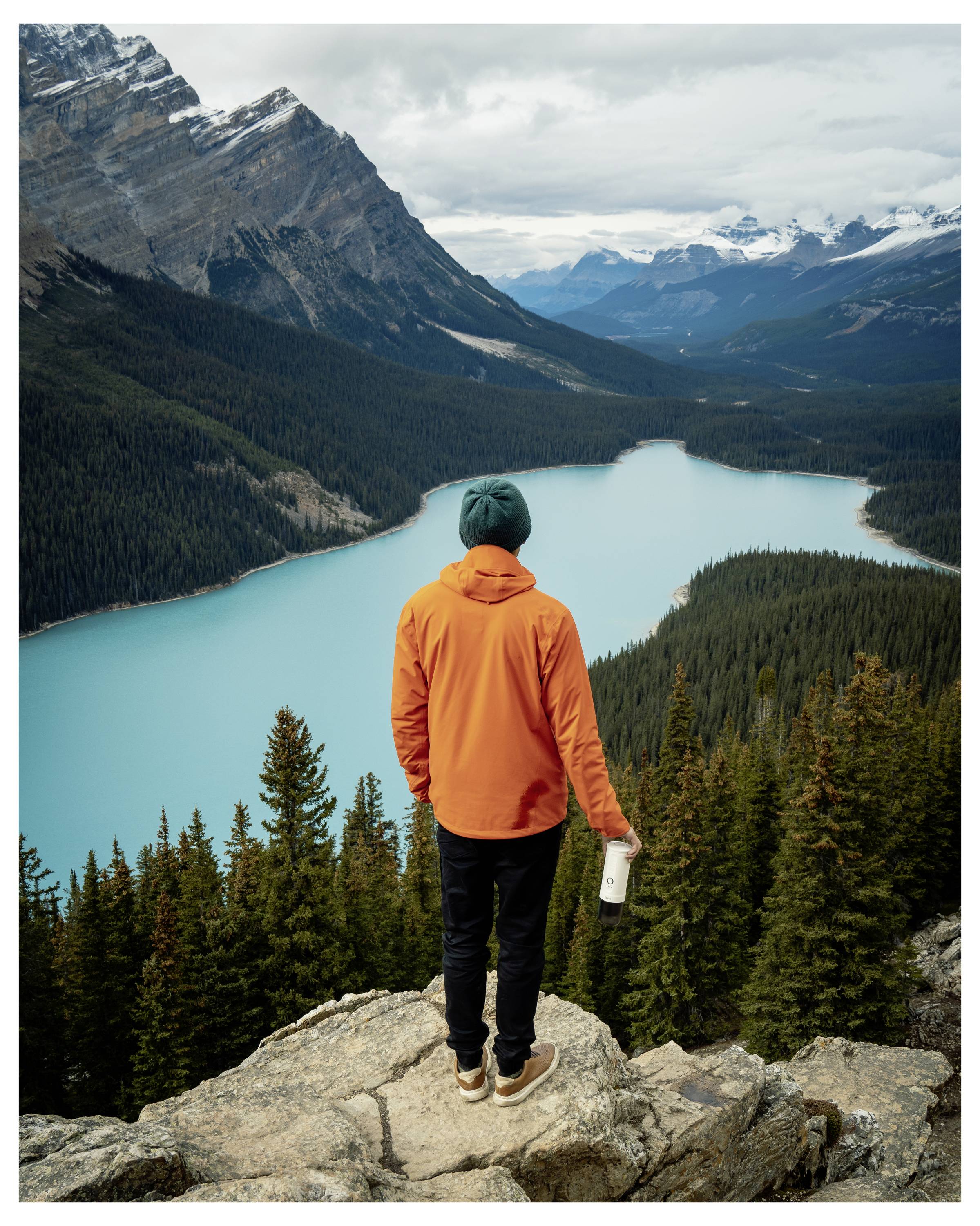 A person in an orange jacket stands on a rocky outcrop overlooking a turquoise lake surrounded by forested mountains under a cloudy sky.