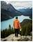A person in an orange jacket stands on a rocky outcrop overlooking a turquoise lake surrounded by forested mountains under a cloudy sky.