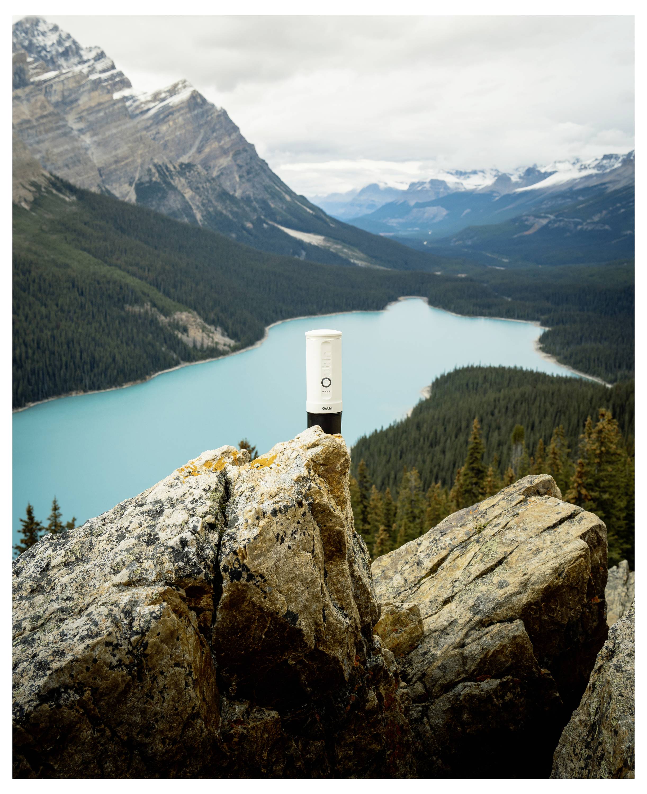 'A cylindrical white object with a logo stands on a rocky cliff overlooking a turquoise lake surrounded by mountains under a cloudy sky.'