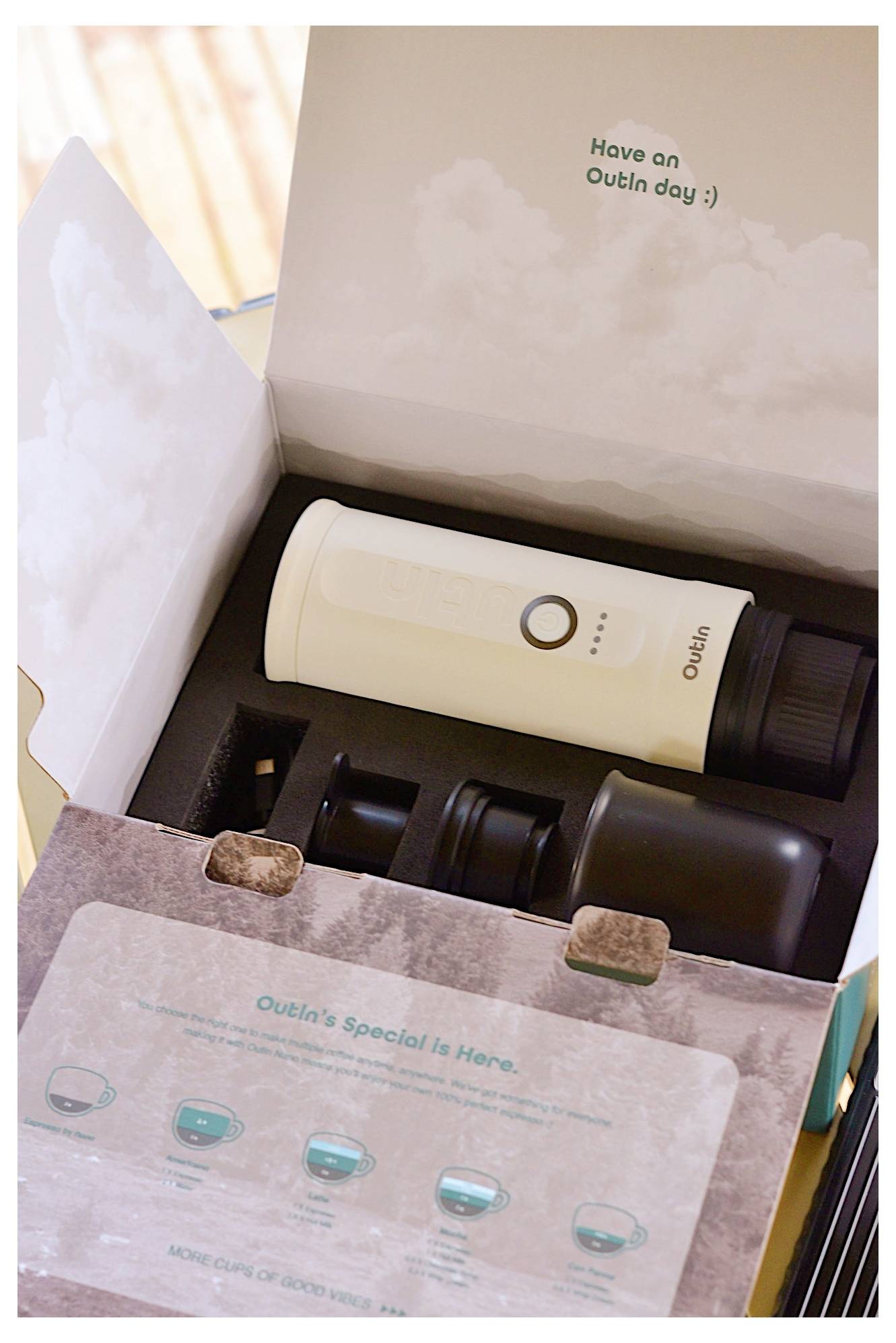 A white portable tumbler and two black lids inside an open cardboard box. The box lid reads 'Have an Outlin day :)'. A leaflet describes product features.