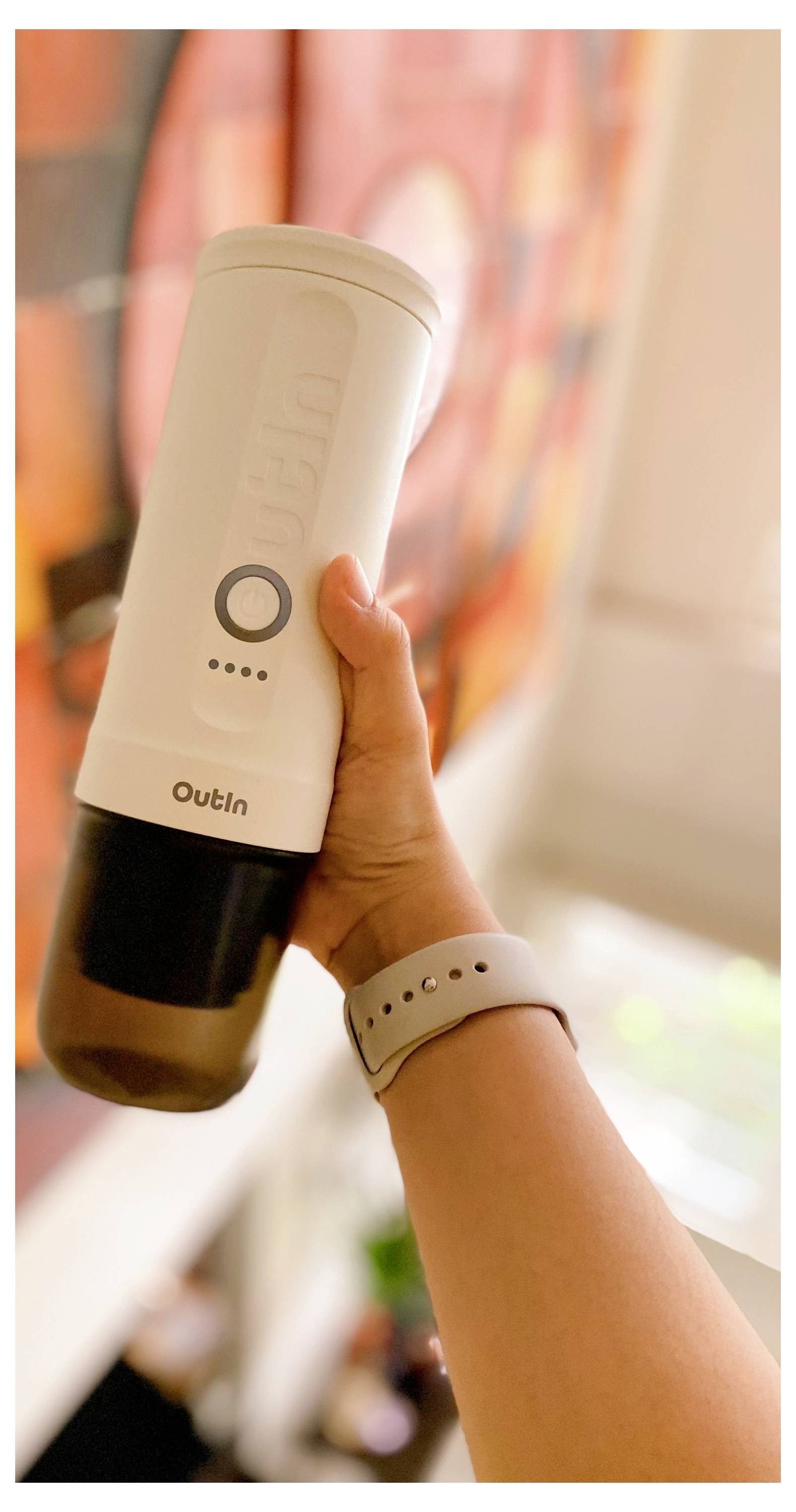 A person holding a portable electric coffee grinder in a well-lit room, with a blurred colorful painting in the background.