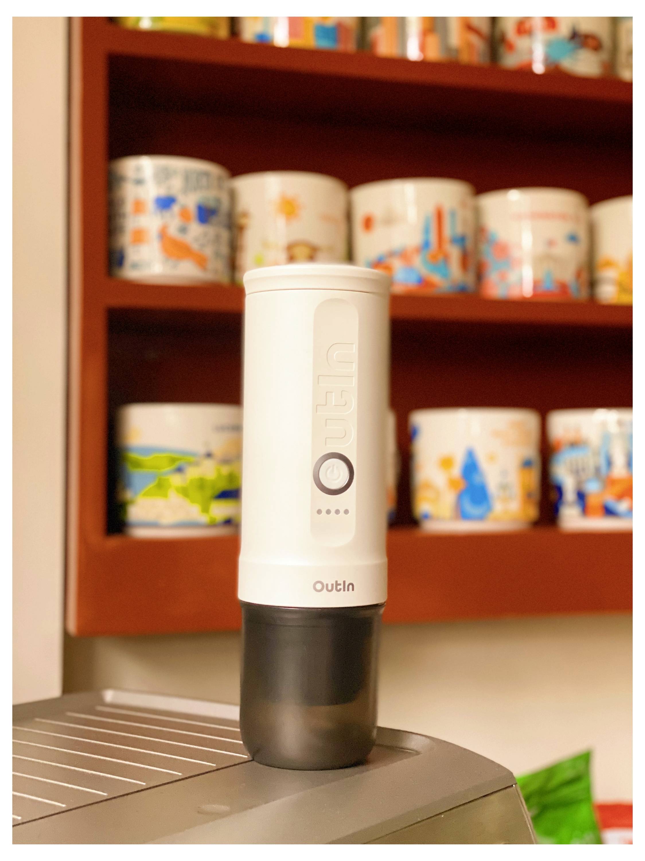 A portable coffee grinder on a kitchen counter, surrounded by colorful mugs on the shelf in the background.