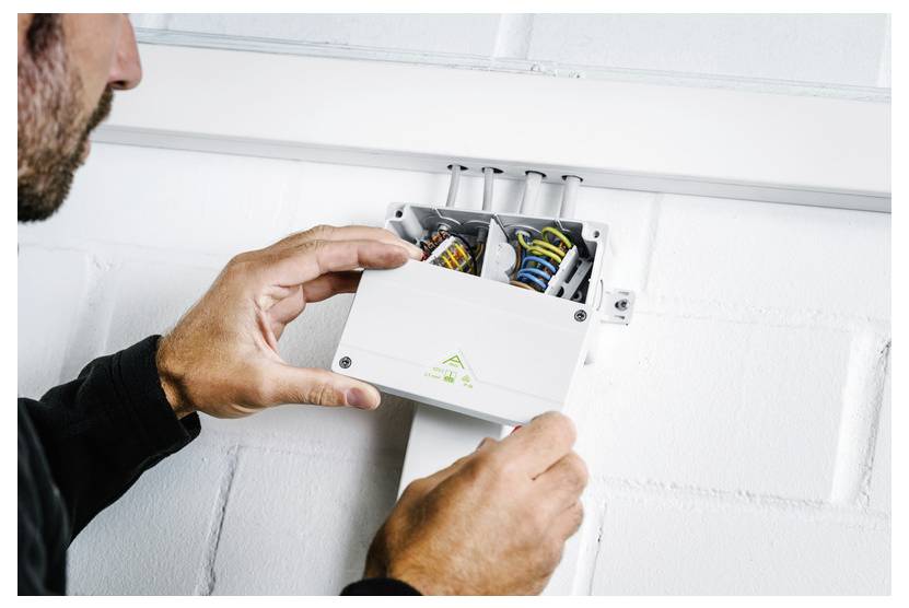 Electrician adjusting wiring inside an open wall-mounted electrical box, with exposed colored cables and conduit above.