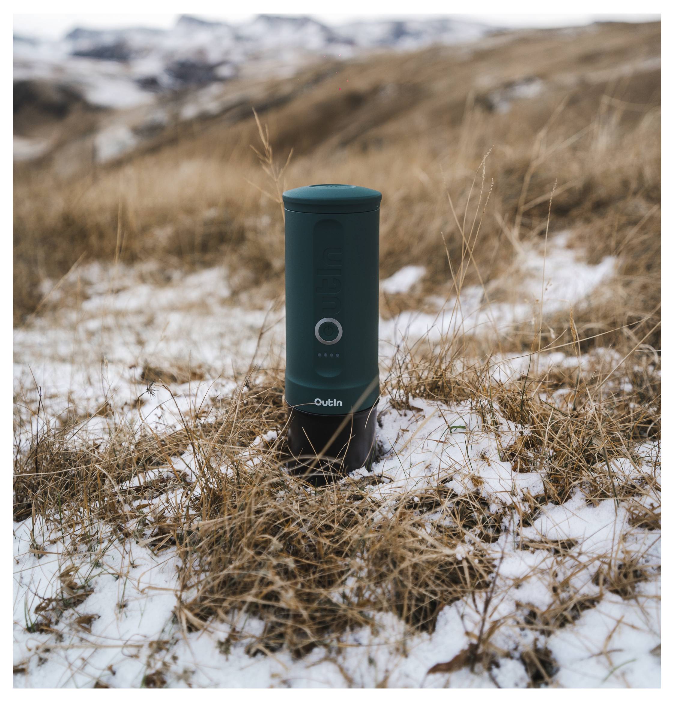 A portable green thermos stands upright on a snowy, grassy field with blurred mountains in the background.