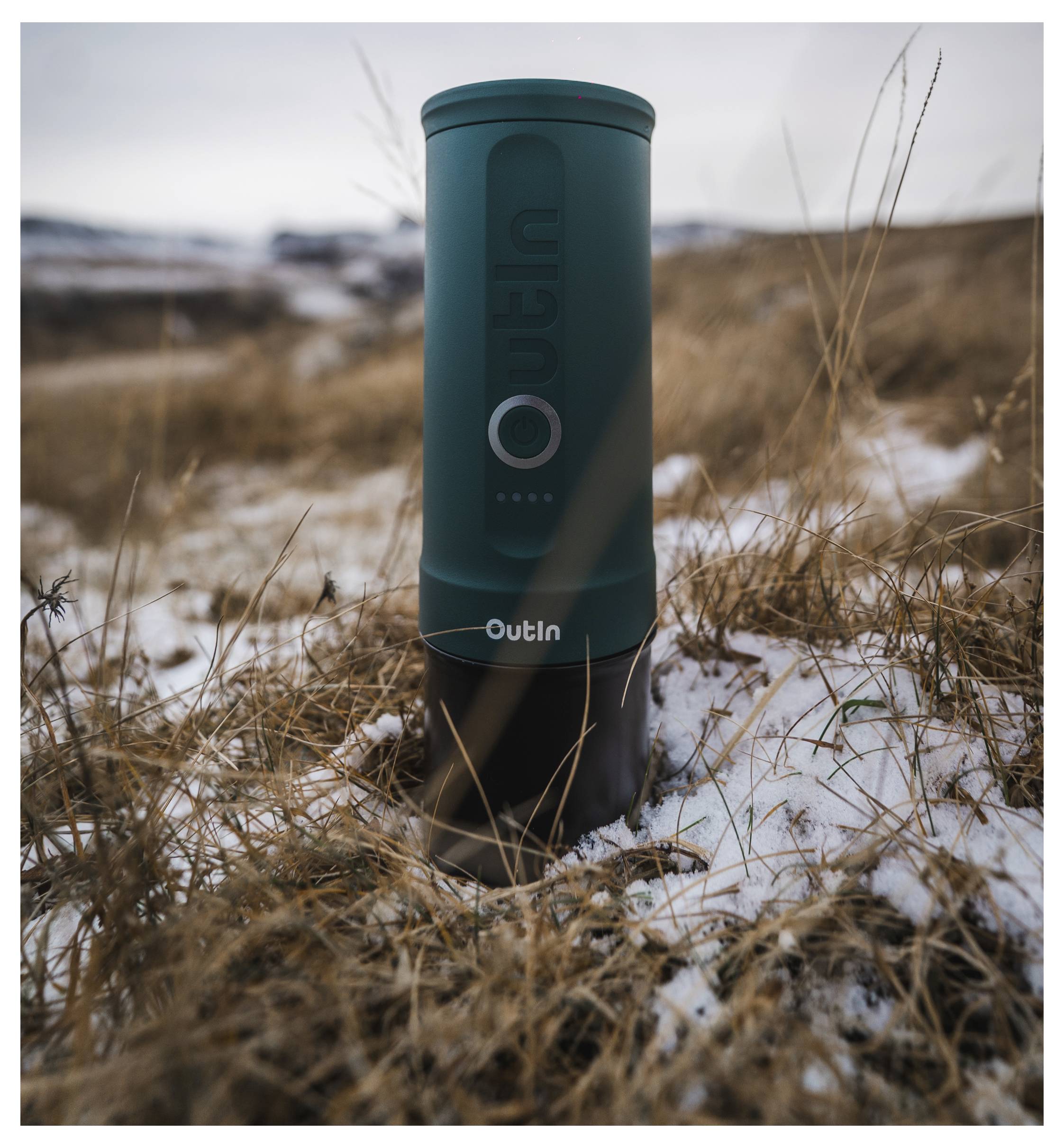 Portable coffee maker standing upright in a snowy, grassy outdoor landscape, highlighting its compact design and rugged use capability.