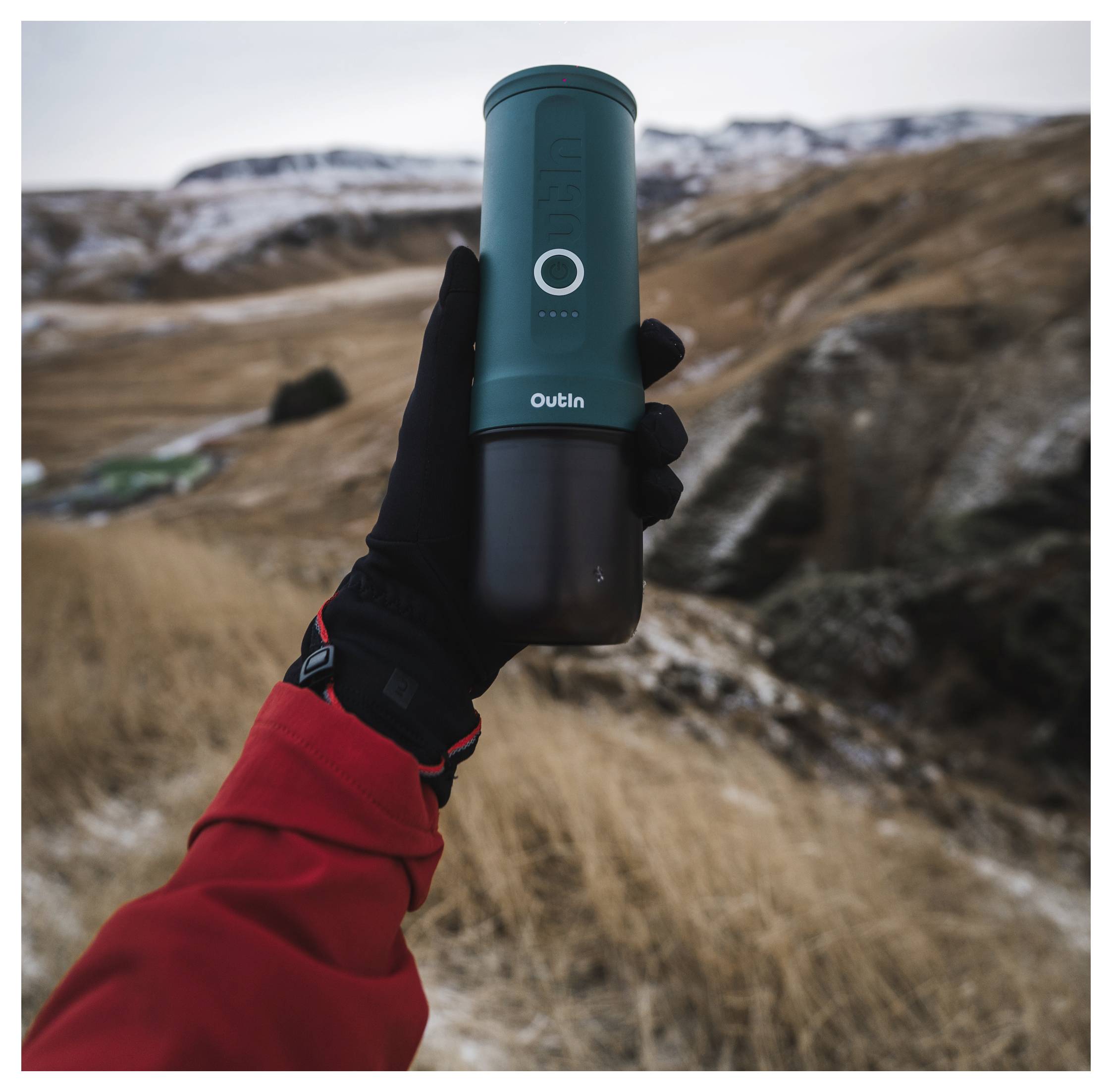 A person in a red jacket and black glove holds a portable thermos in a mountainous landscape, signifying outdoor adventure.