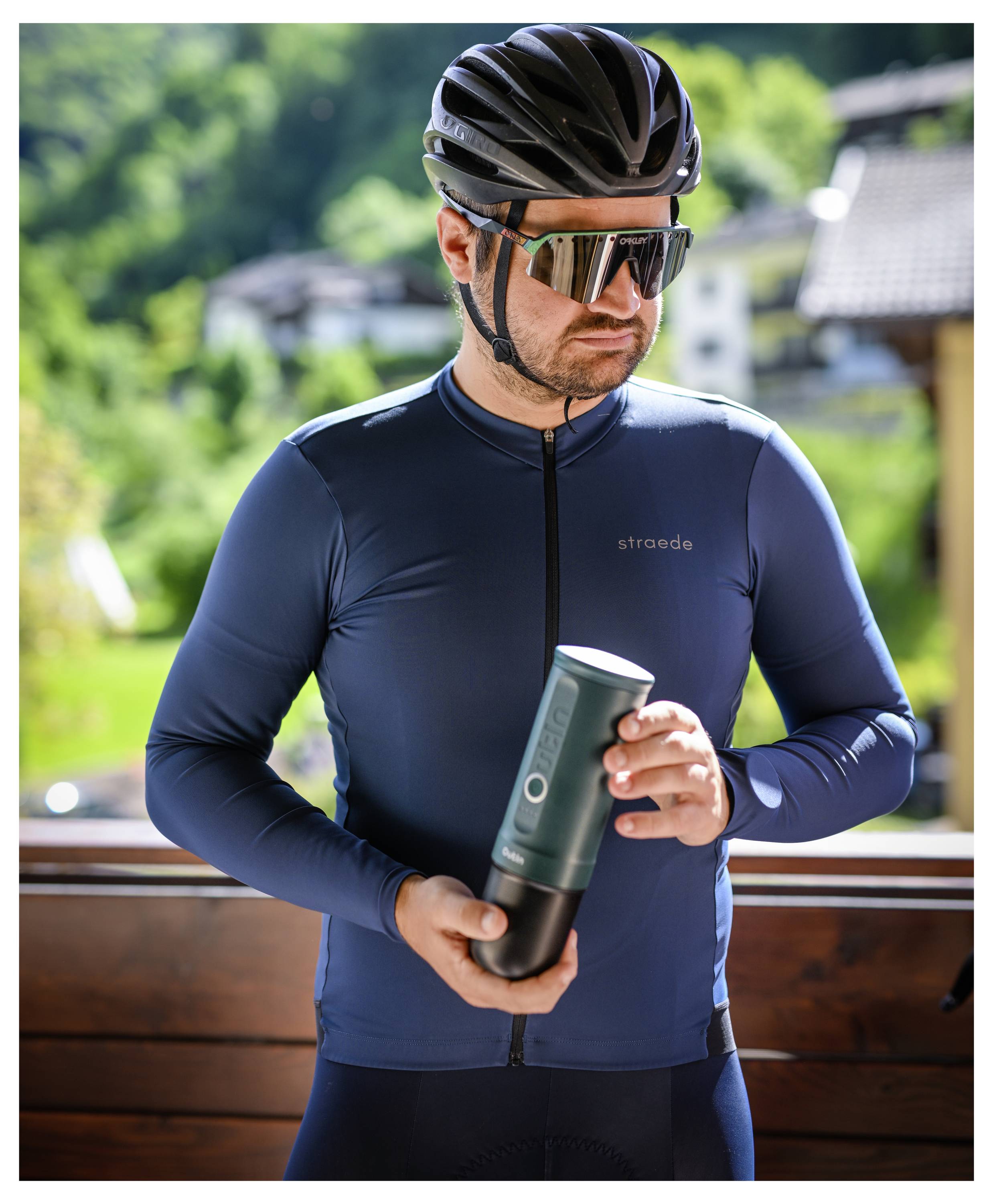 A cyclist wearing sunglasses and a helmet holds a cycling bottle outdoors, with houses and greenery in the background.