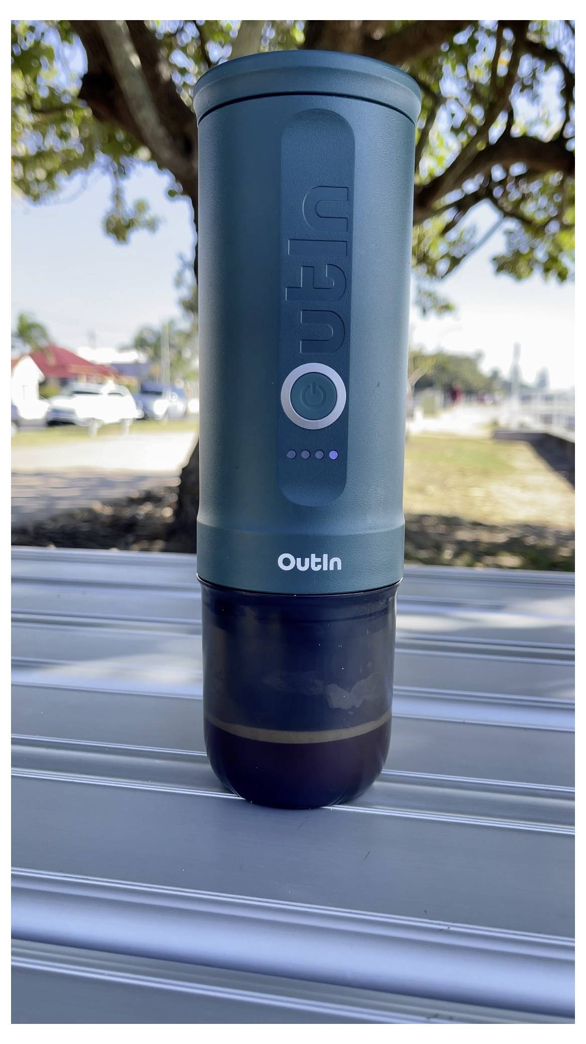 A teal portable coffee maker labeled 'Outin' sits on a metal surface outdoors, with a tree and suburban street in the background.