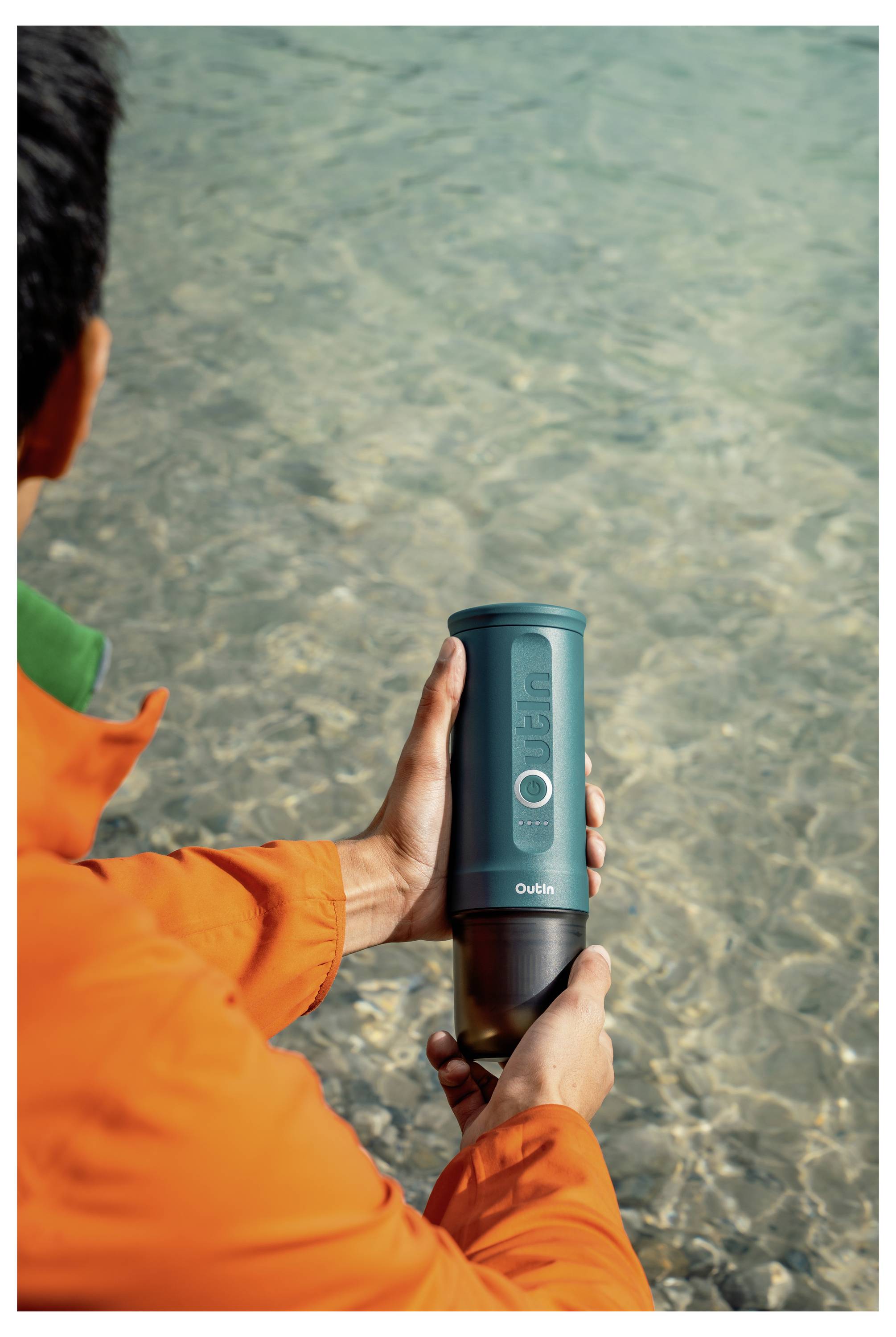 A person in an orange jacket holds a portable insulated bottle with a green exterior above clear water.