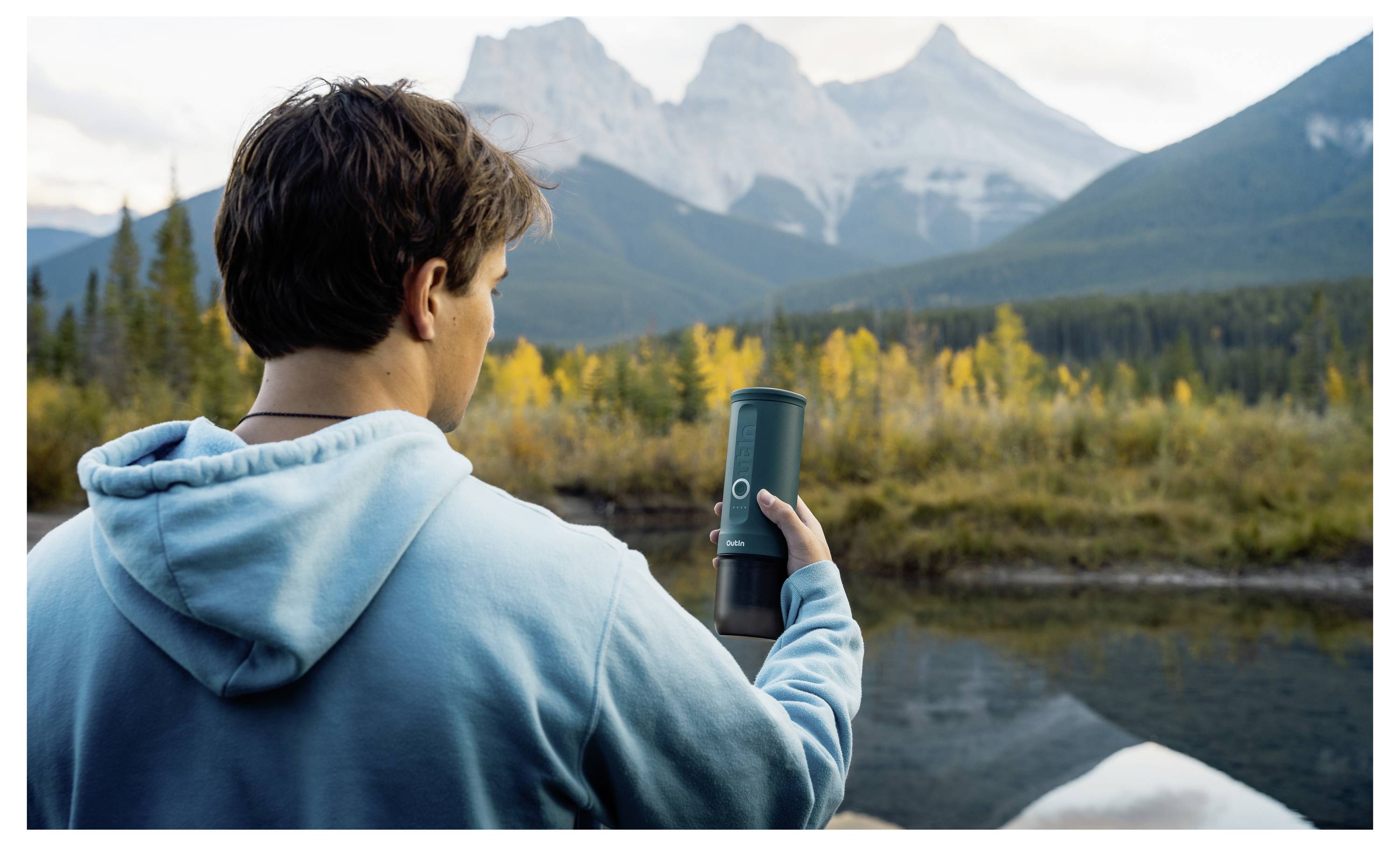 A person in a blue hoodie holds a tumbler, gazing at mountain peaks across a forested landscape with a reflective body of water.