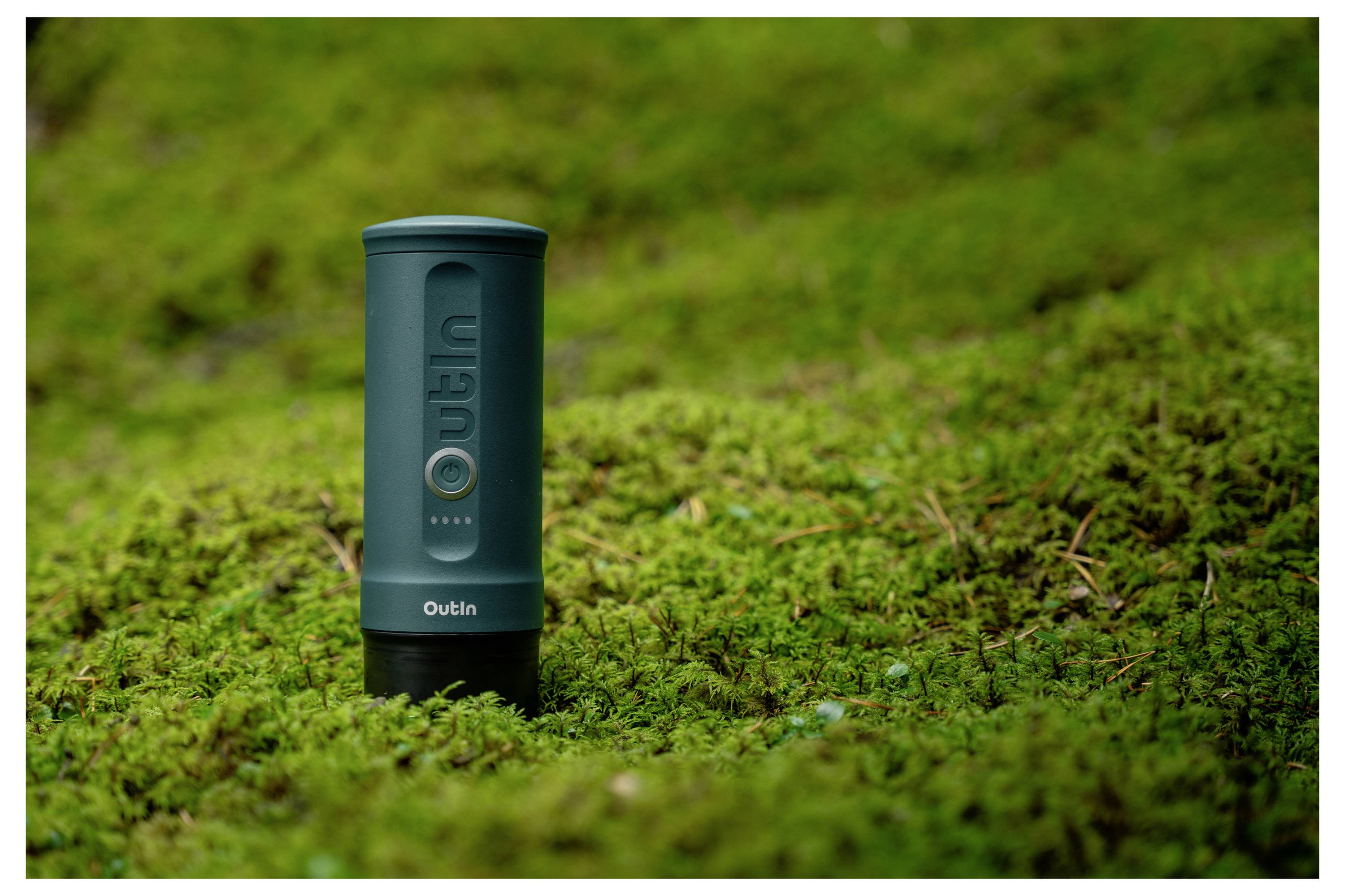 A portable, cylindrical speaker with the brand 'Outin' stands on a lush, green moss-covered surface, blending with the natural setting.