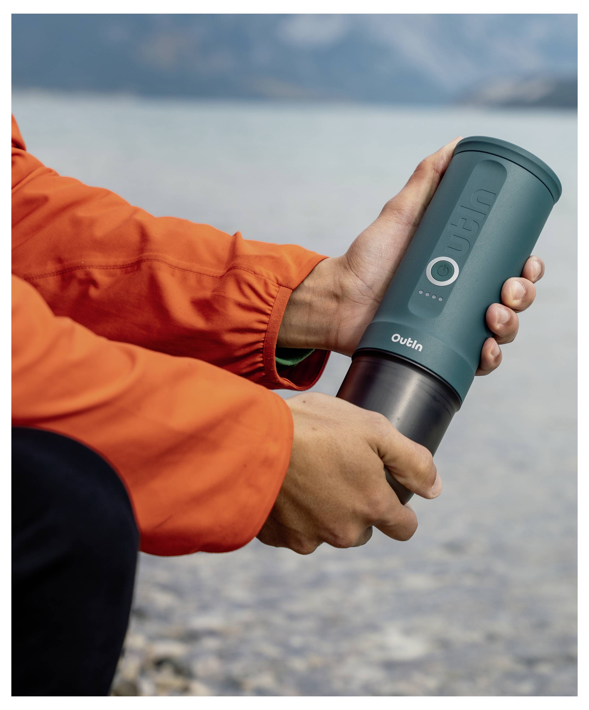 A person wearing an orange jacket holds a green portable coffee maker by a lake, emphasizing outdoor use and convenience.