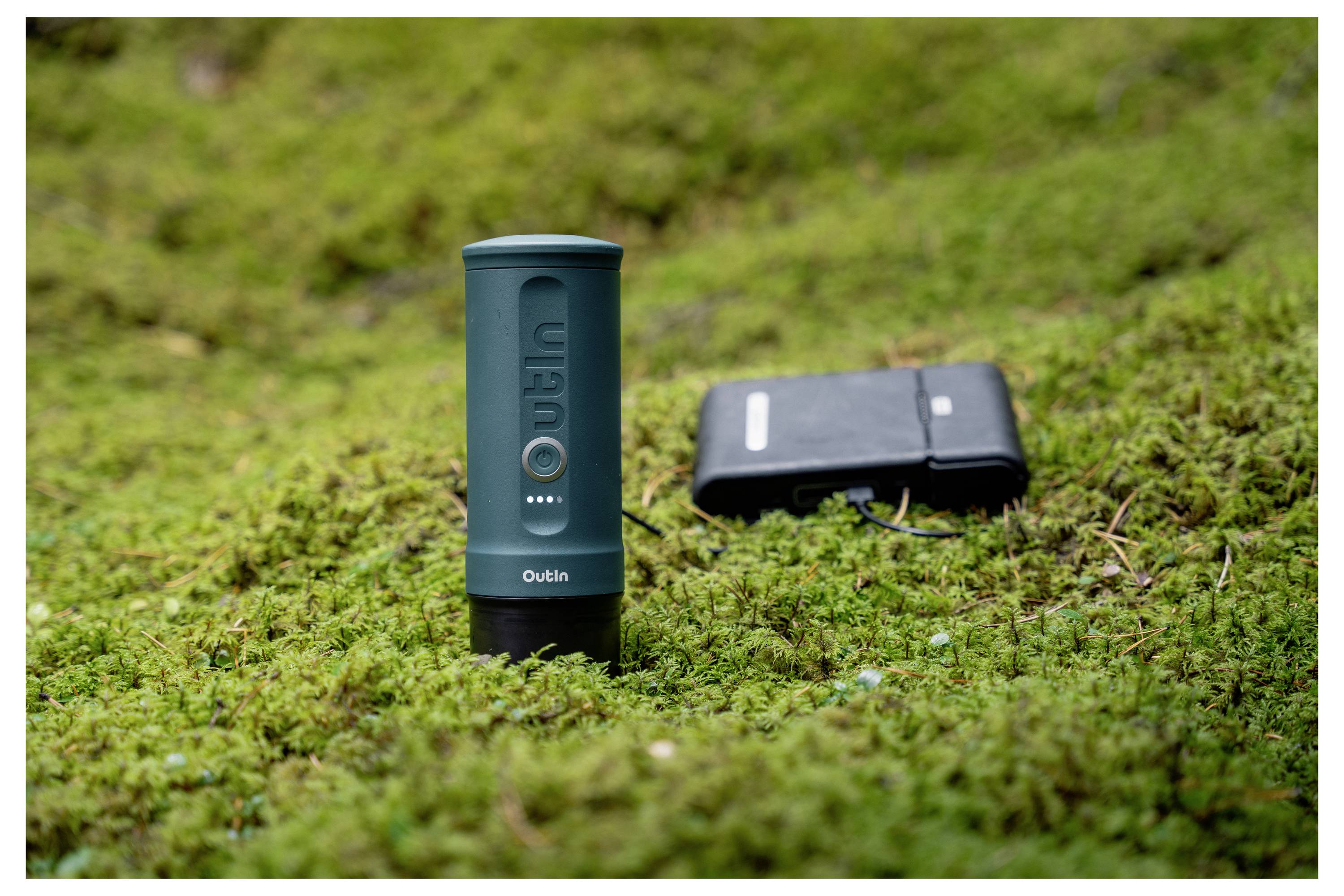 A portable power bank and a compact camping stove are placed on green moss, suggesting outdoor use and convenience for travel.