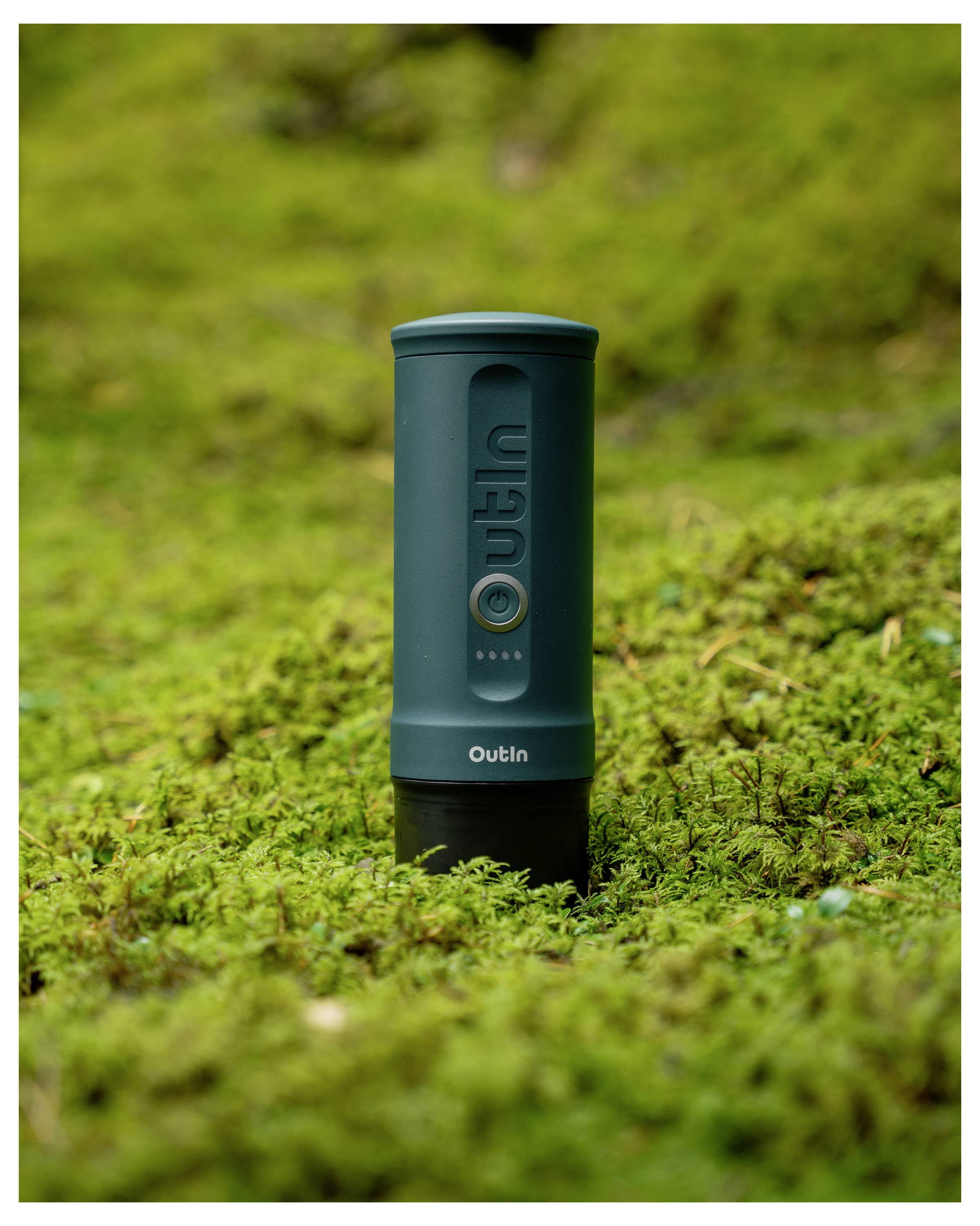 A cylindrical, dark green coffee container labeled 'Outin' stands upright on a mossy forest floor, blending with the natural surroundings.