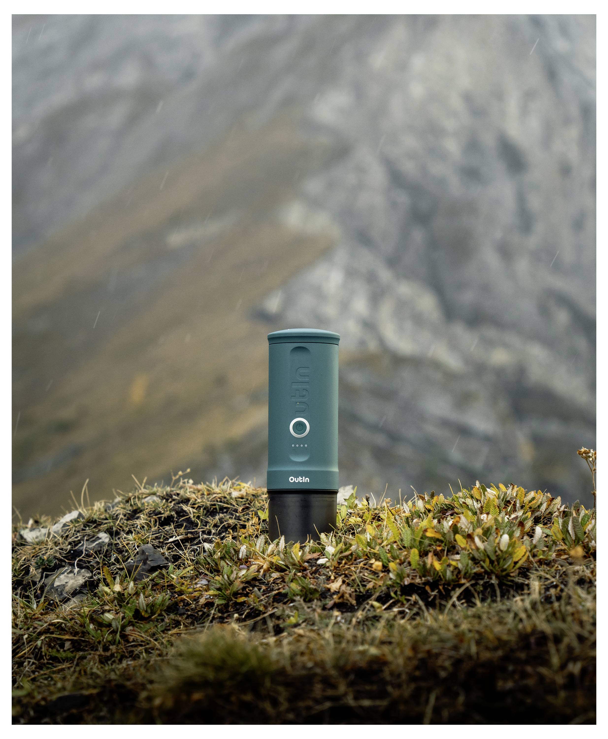 A green travel mug stands on rocky terrain with mountains in the background, suggesting an outdoor adventure setting.