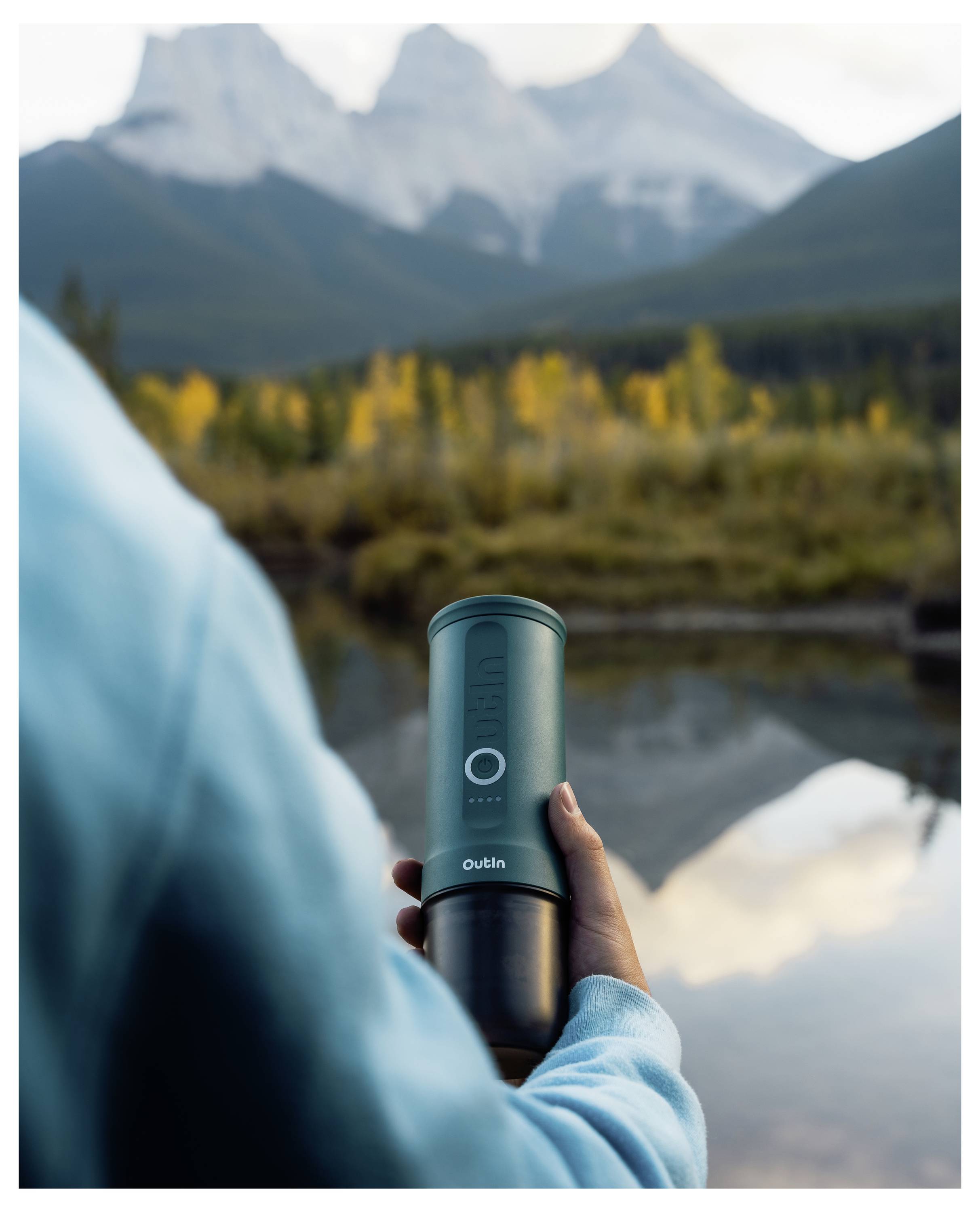 Person in blue holds a portable coffee grinder by a serene lake, with vibrant autumn trees and distant mountains in the background.