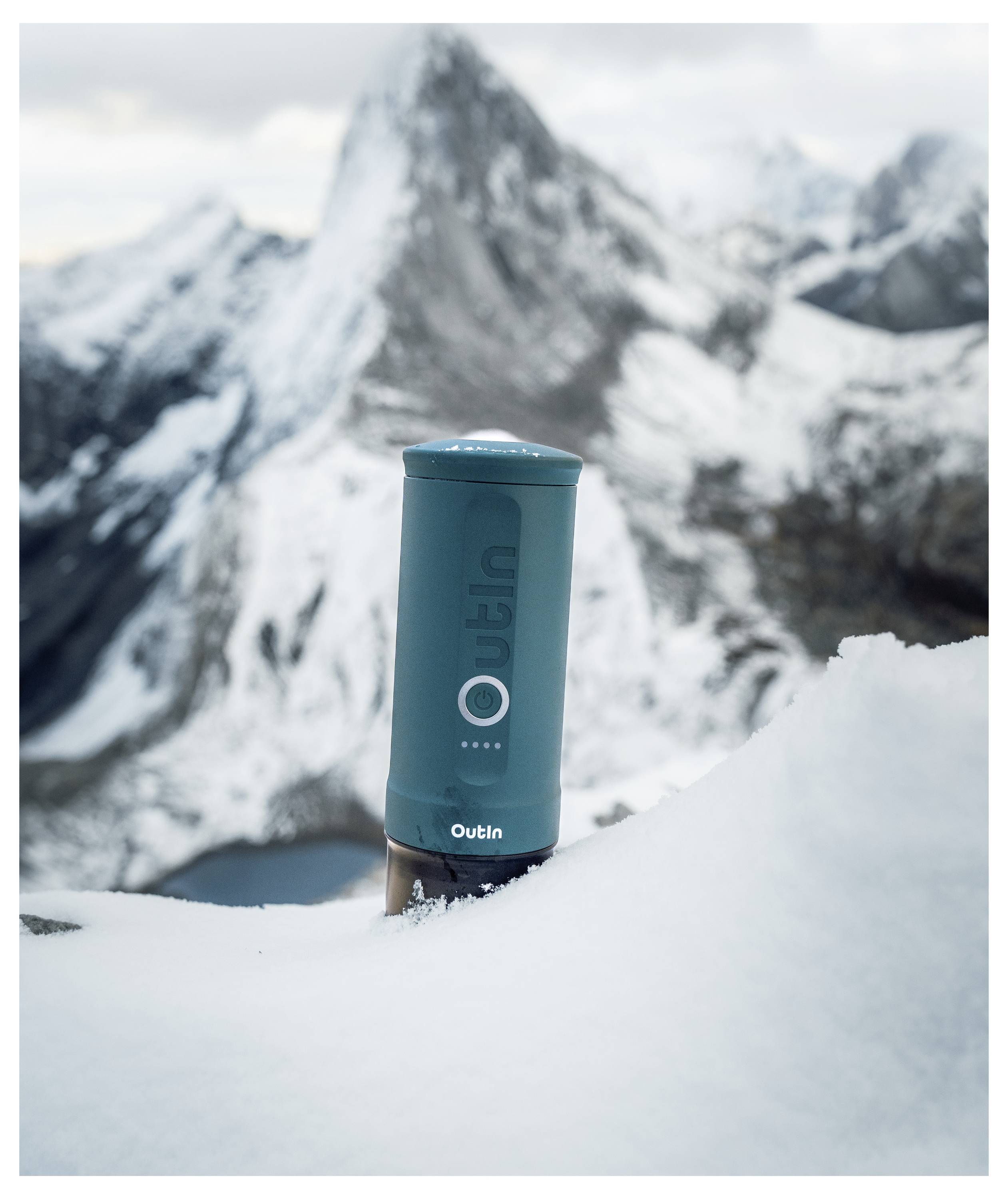 A blue Outin branded thermos is partially buried in snow with a backdrop of snow-covered mountains under a cloudy sky.